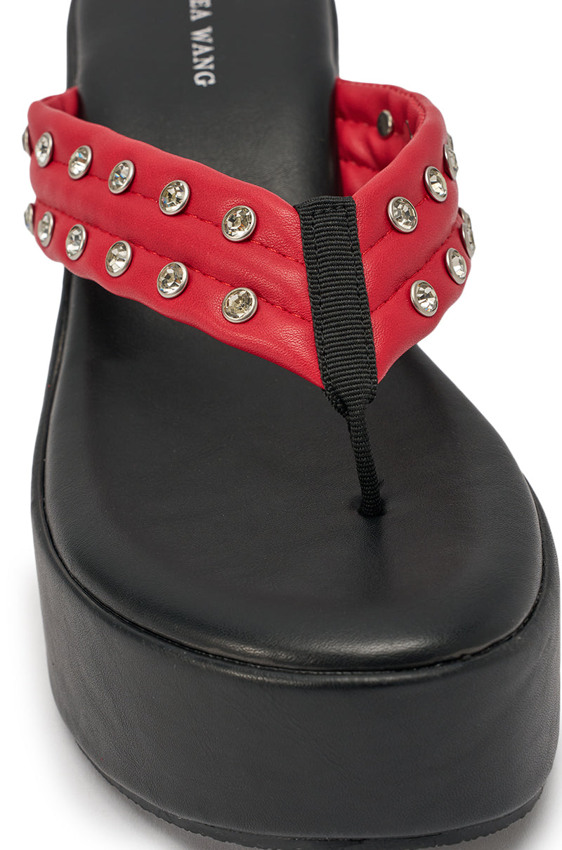 The AZALEA WANG NOVELLE BLACK AND RED STUD EMBELLISHED PLATFORM SANDAL features a black wedge sole, padded red thong strap with silver studs and clear rhinestones, and visible brand text on the footbed. 6