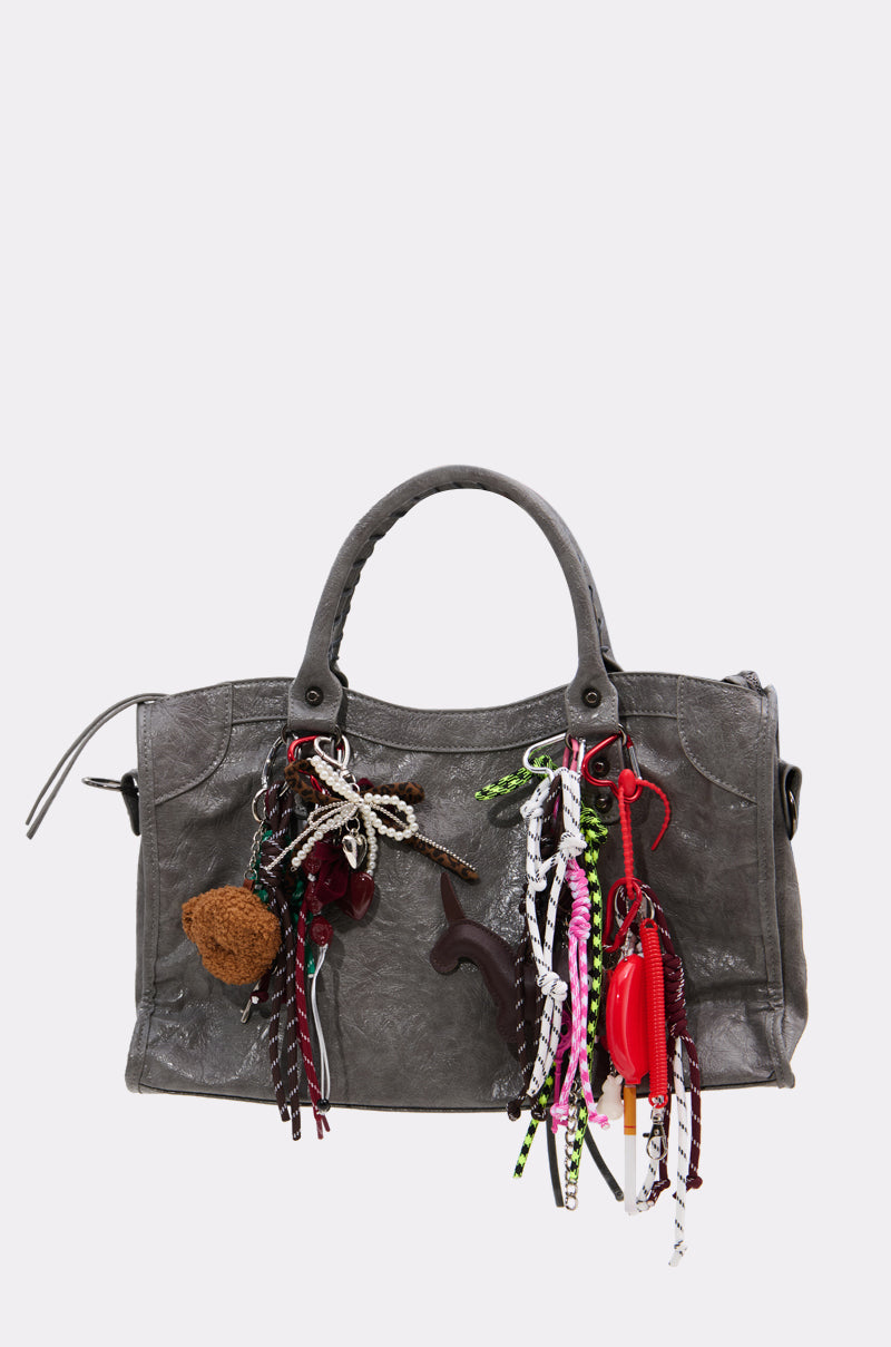 A gray leather handbag decorated with the ULTIMATE KEYCHAIN SET—featuring colorful novelty charms, beads, a fuzzy brown pom-pom, red and white cords, and other assorted decorative items hanging from the handles.