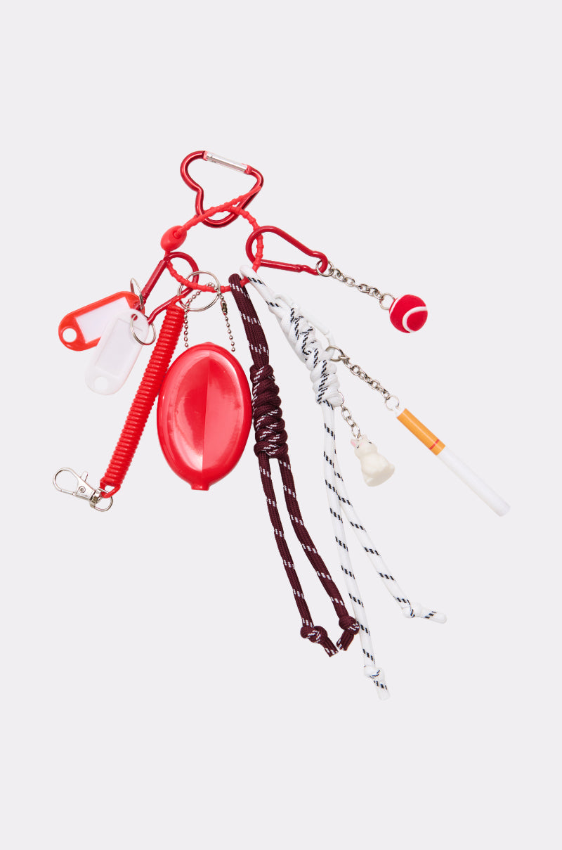 The ULTIMATE KEYCHAIN SET is a bold accessory featuring red, white, and brown novelty keychains—carabiner, pill capsule, tennis ball, whistle, coiled cord, football, rope knots, and cigarette—displayed on a light background.