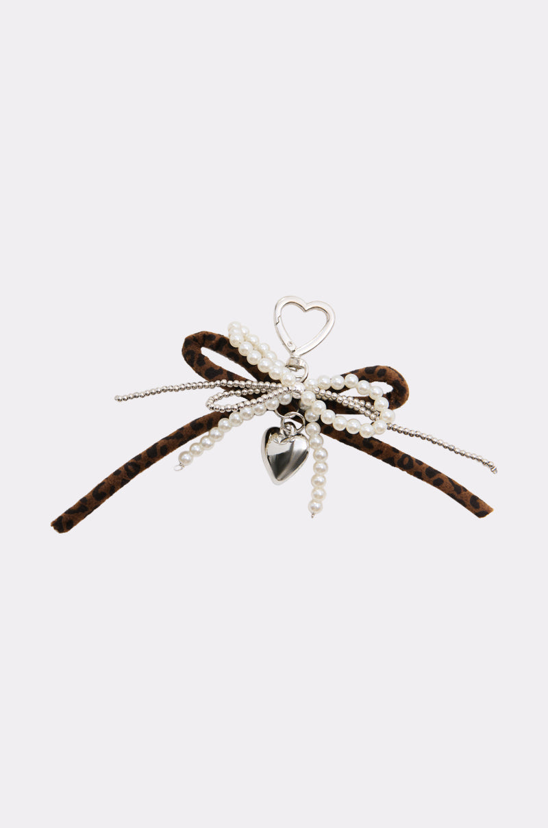A bold accessory, the ULTIMATE KEYCHAIN SET features a leopard print ribbon, faux pearl strands, silver heart-shaped outline, and small heart charm arranged in a bow shape on a white background.