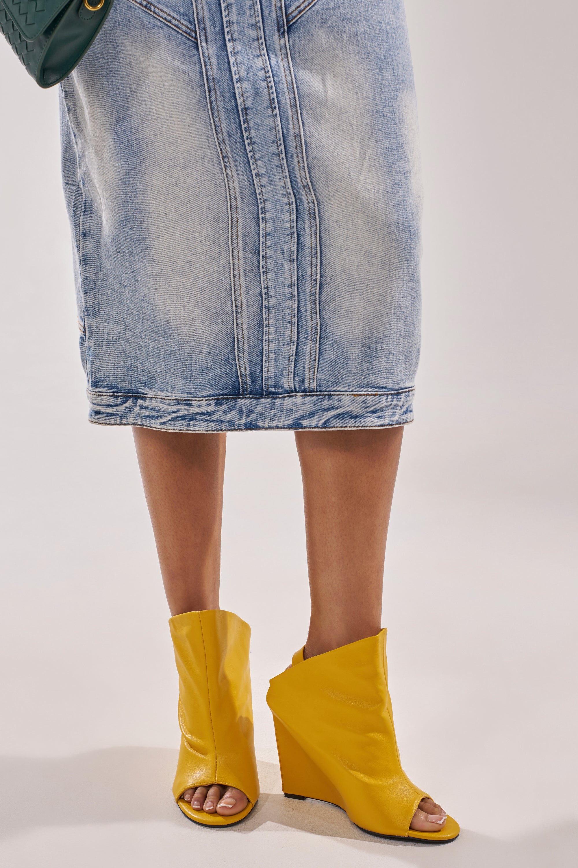 A person wears a knee-length light blue denim skirt and AZALEA WANG NOVIS YELLOW WEDGE BOOTIE almond peep toe boots, standing on a light background. 1