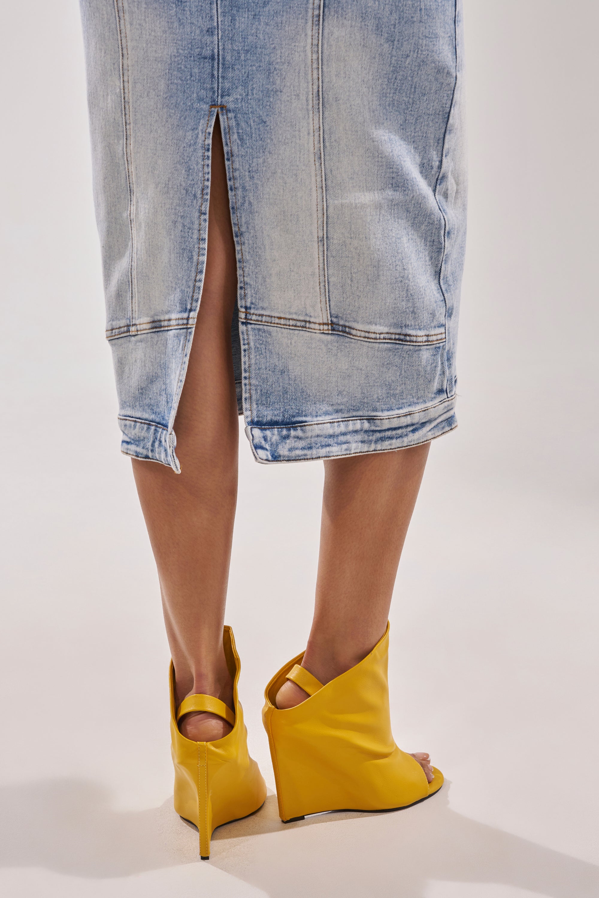 A person stands, shown from behind against a plain background, wearing bright AZALEA WANG NOVIS YELLOW WEDGE BOOTIE shoes and a light blue denim skirt with a back slit. 2