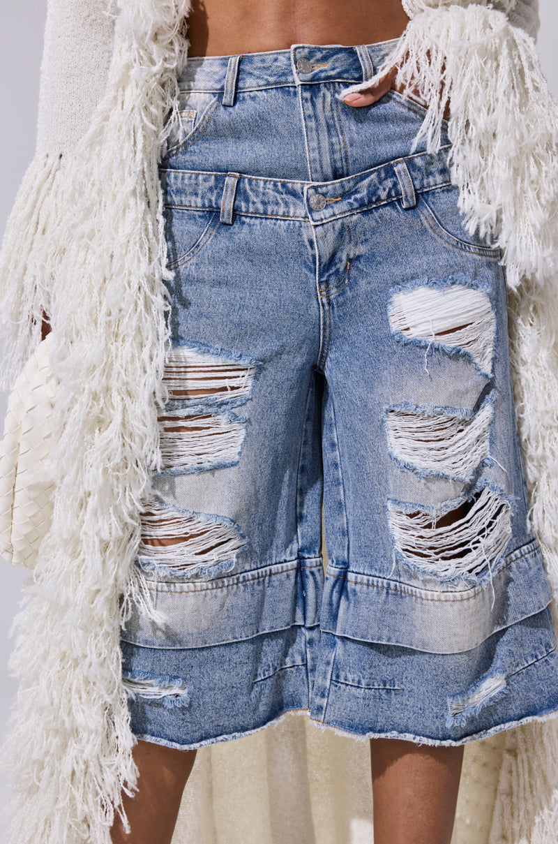 A person wears the GO GET SOME DENIM BERMUDA SHORT—distressed, high-rise denim with frayed holes—paired with a textured, fringed white jacket. The bold outfit highlights the shorts’ rugged blue look. 0