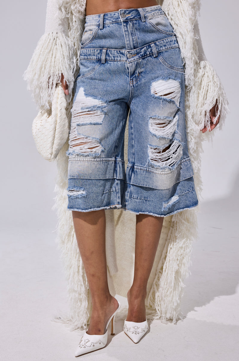 Wearing the GO GET SOME DENIM BERMUDA SHORT, a long textured white cardigan, white pointed-toe heels, and holding a white woven clutch. 2