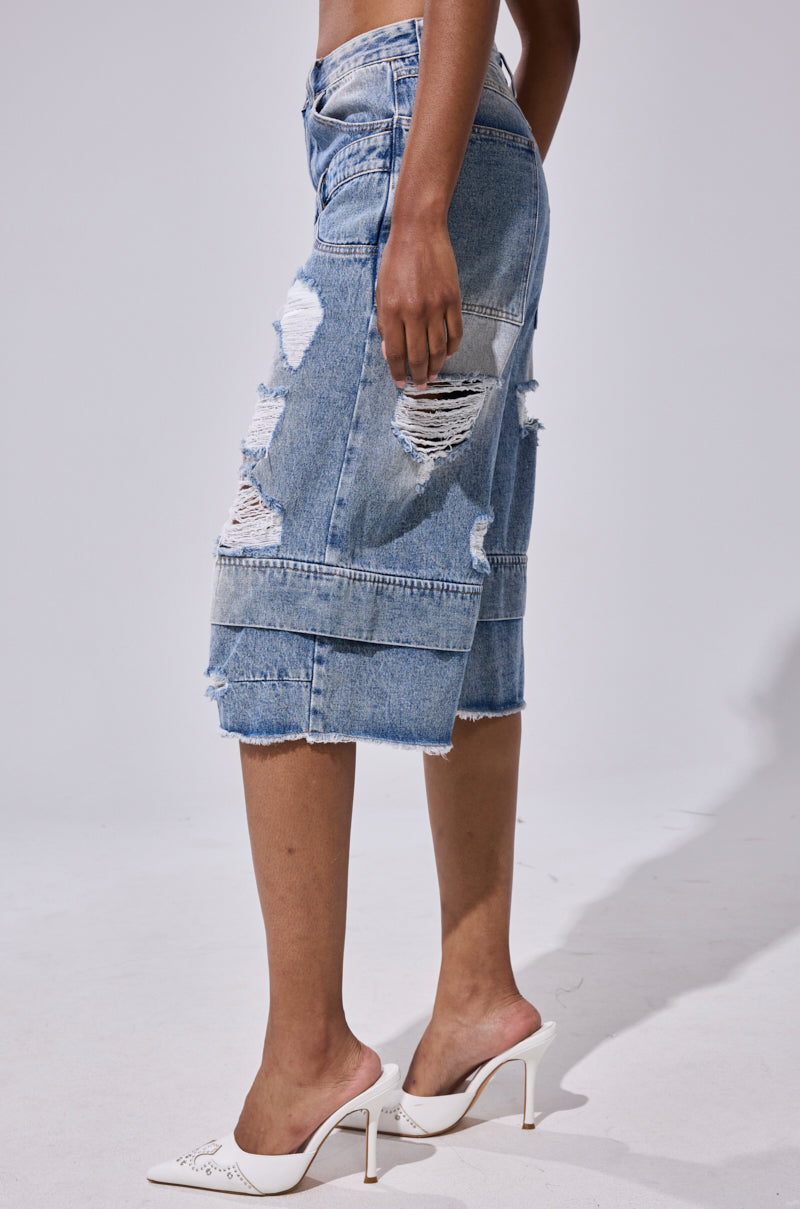 A person wearing the GO GET SOME DENIM BERMUDA SHORT, featuring distressed, ripped detailing, and white heeled mules stands against a plain light background. 3