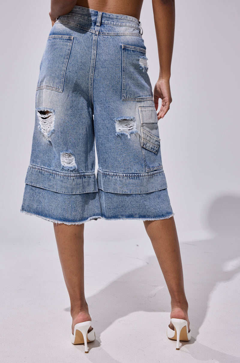 A person wears the GO GET SOME DENIM BERMUDA SHORT, featuring distressed denim, frayed hems, a high-rise fit, and large back pockets, paired with white high-heeled shoes against a plain background. 4