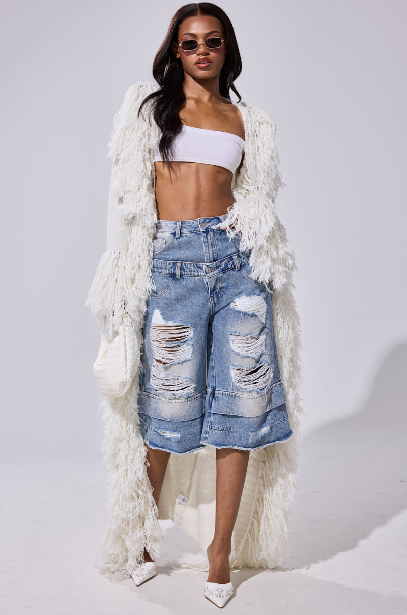 A woman wearing sunglasses, a white bandeau, textured coat, white heels, and holding a woven bag walks confidently in GO GET SOME DENIM BERMUDA SHORTS with ultra-distressed details against a plain background. 6