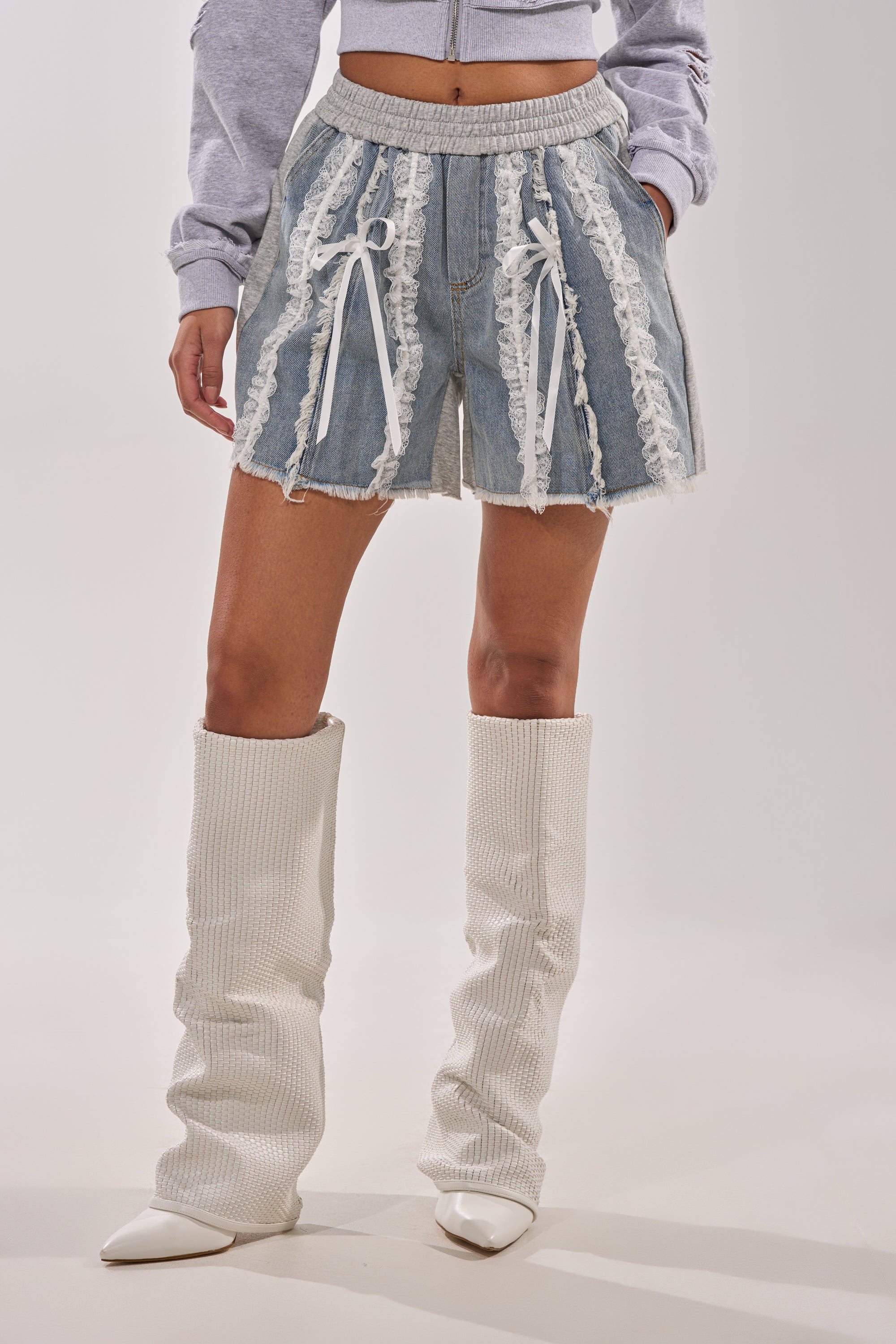 A person wears the TIED AND LACED DENIM SHORTS with white lace accents and bows, paired with a gray cropped jacket and tall white textured pointed-toe boots. The background is plain and light-colored. 1