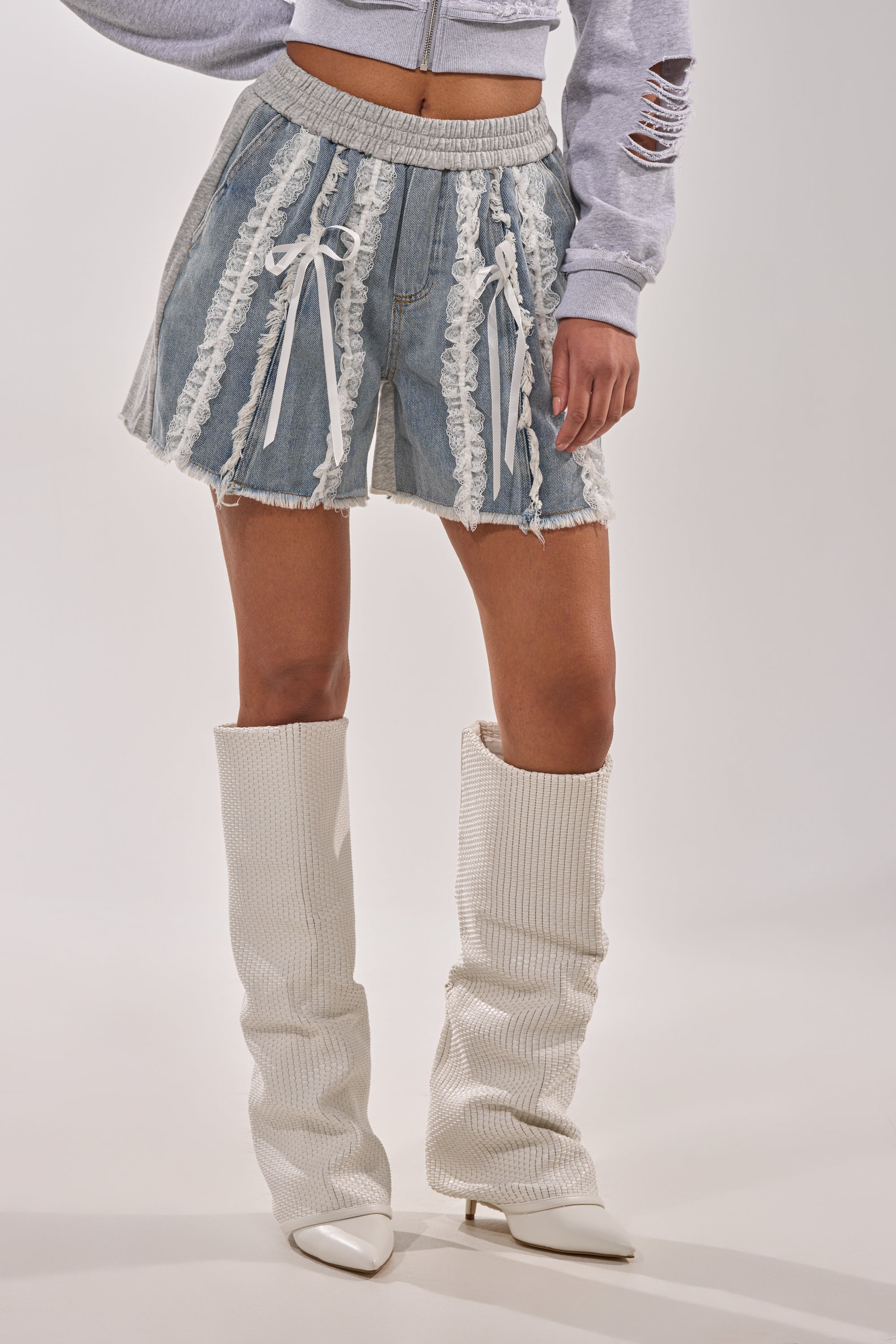 A person wearing TIED AND LACED DENIM SHORTS, a cropped gray sweatshirt, and white textured pointy-toe knee-high boots stands against a plain background. 2