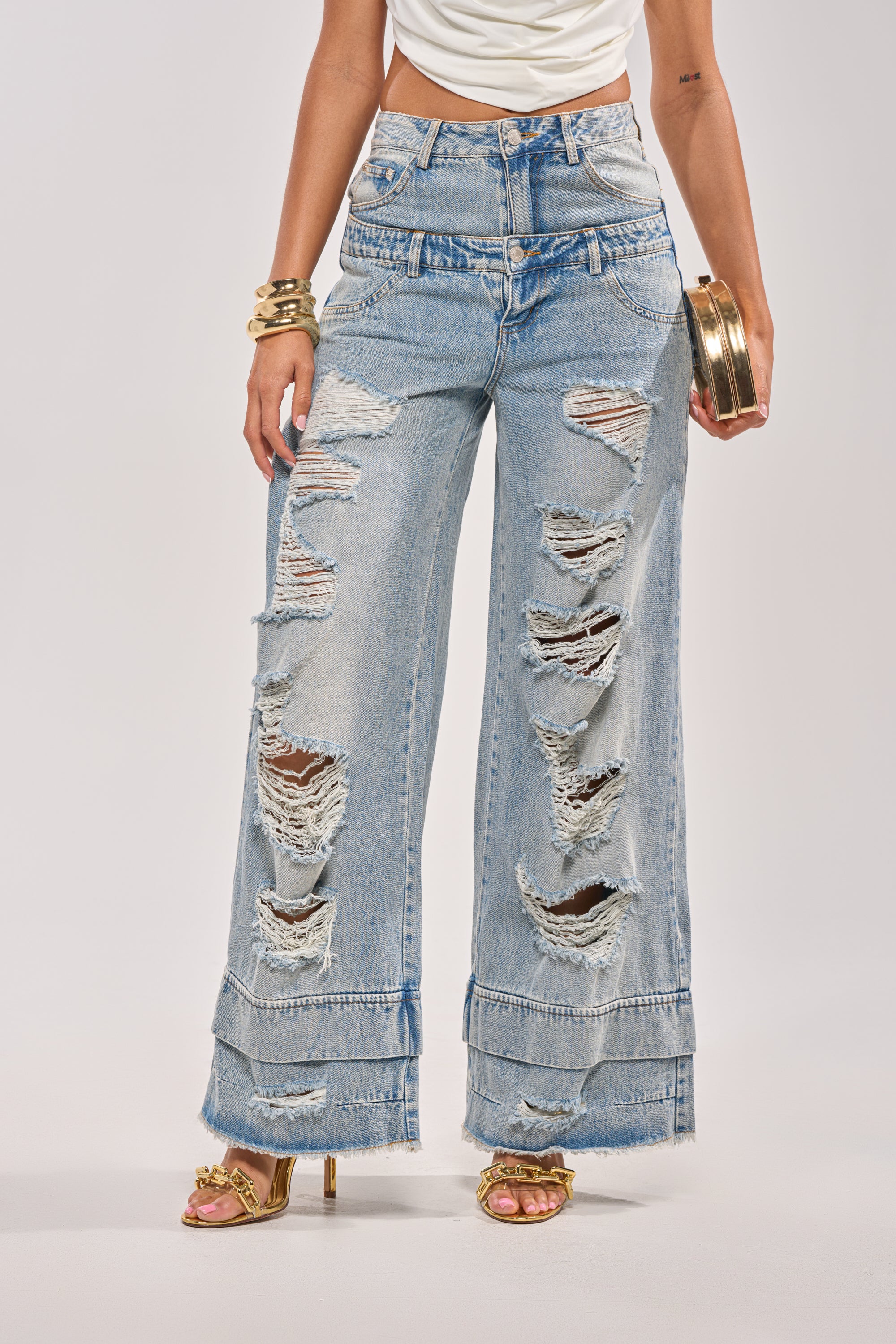 A person wearing GO GET SOME DOUBLE WAISTED DENIM, gold heeled sandals, a white top, gold bracelets, and holding a small round gold handbag. 0
