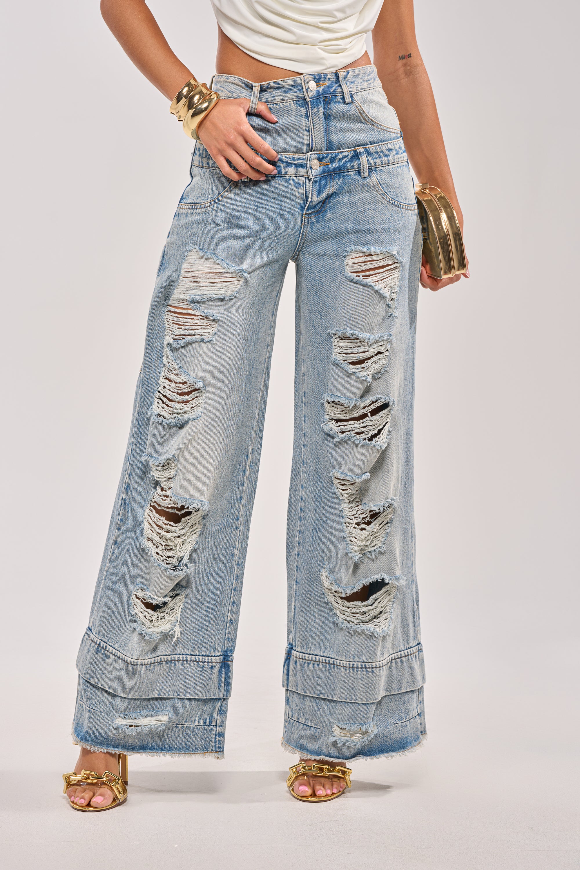 Someone stands against a plain background wearing GO GET SOME DOUBLE WAISTED DENIM—light blue, high-rise wide leg distressed jeans with large rips—paired with gold high-heeled sandals and chunky gold bracelets. 2