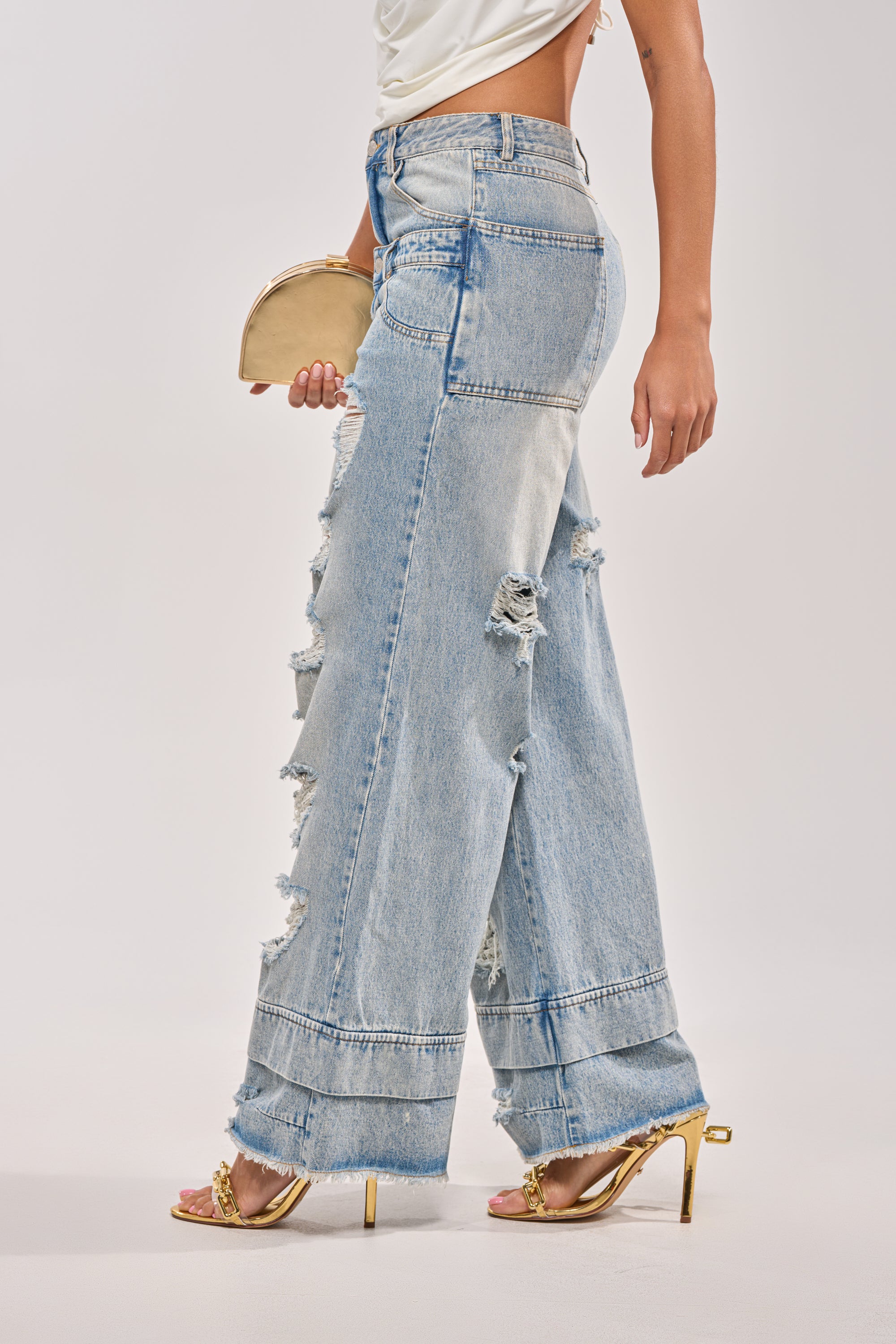 Wearing the GO GET SOME DOUBLE WAISTED DENIM, a white top, gold heels, and holding a beige clutch, a person poses against a plain light background. 3