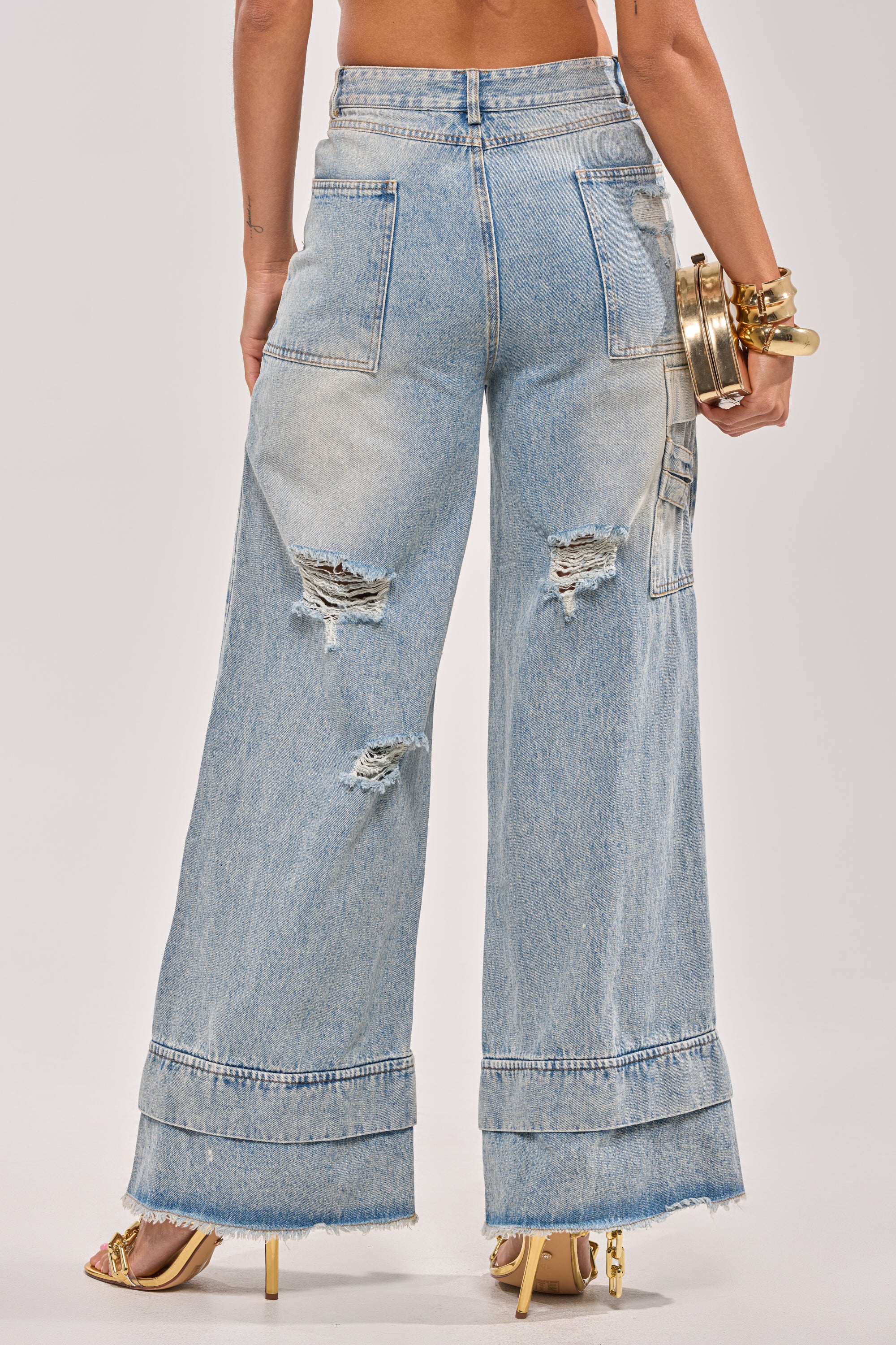 Someone wearing the GO GET SOME DOUBLE WAISTED DENIM—high-rise wide-leg jeans with distressed patches and raw hems—paired with heeled sandals, gold bracelets, and a gold clutch. The photo captures the outfit from the back. 4
