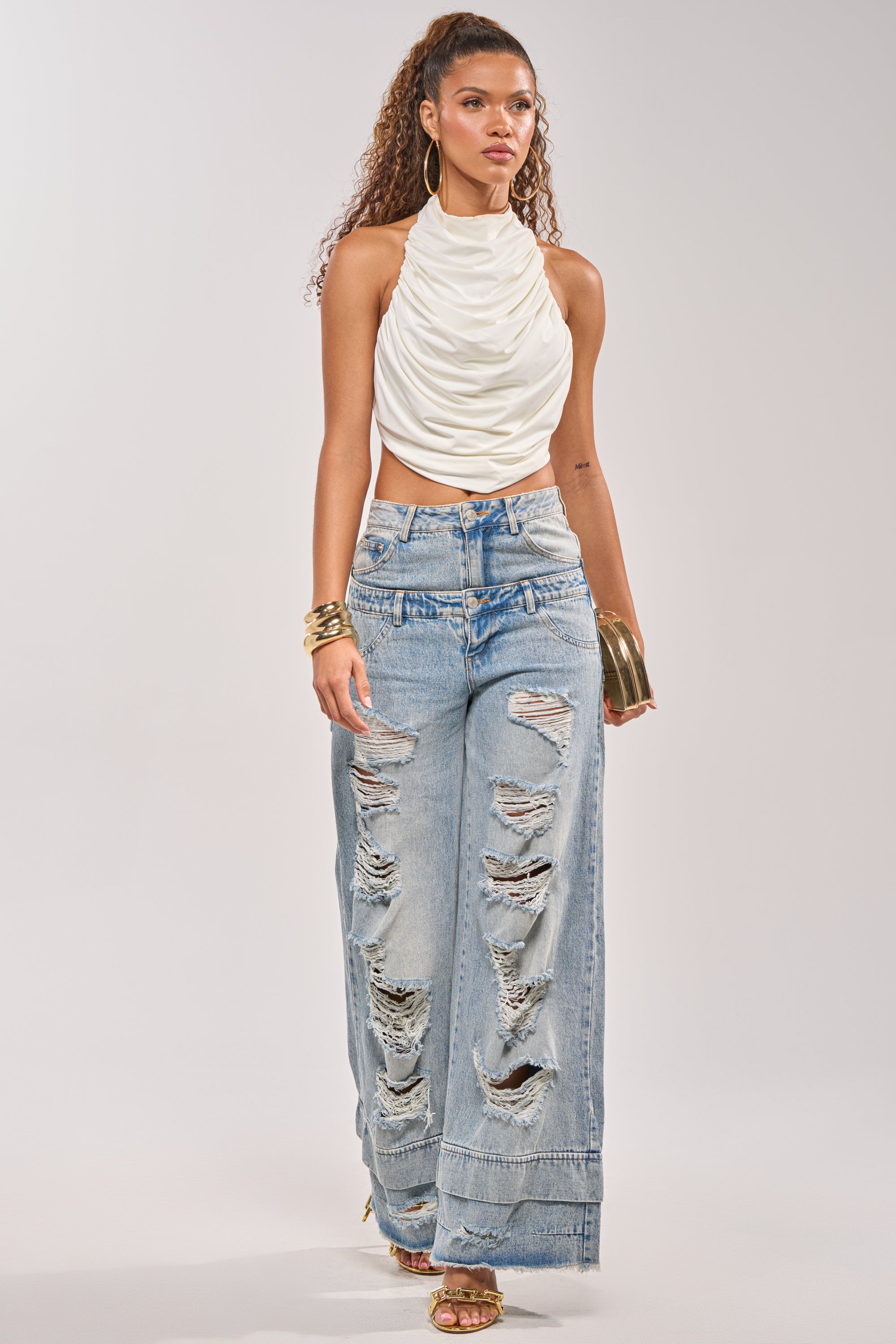 A woman models the GO GET SOME DOUBLE WAISTED DENIM—high-rise wide-leg jeans with heavy rips—paired with a white sleeveless halter top, gold hoop earrings, a wide gold bracelet, and heeled sandals against a plain background. 6