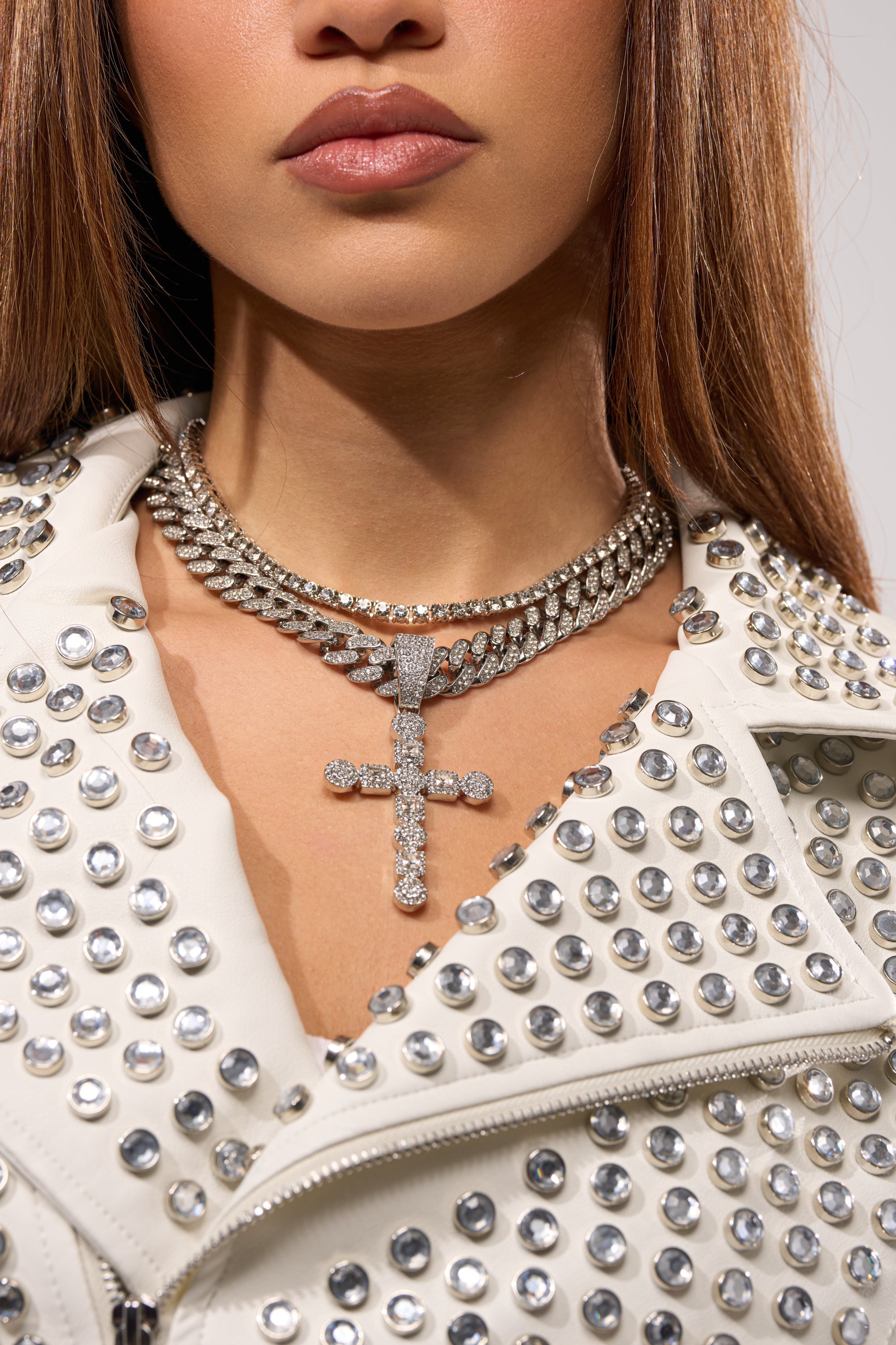 A woman wears a white studded leather jacket and the WHAT U SAY NECKLACE, featuring pavé Cuban link chains and a bold rhinestone cross pendant. Only the lower half of her face and neck are visible. 5