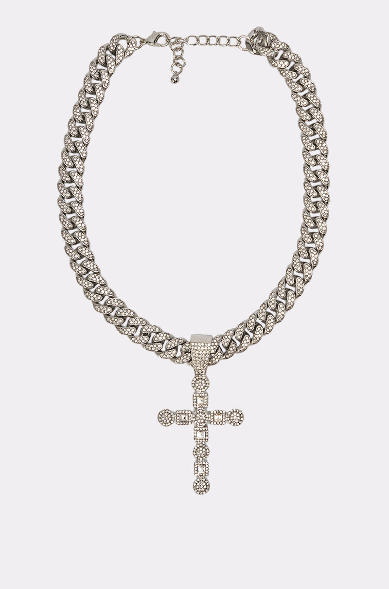 The WHAT U SAY NECKLACE features a thick silver Cuban link chain set with diamonds and a large sparkling cross pendant adorned with small rhinestones, displayed on a simple white background. 2