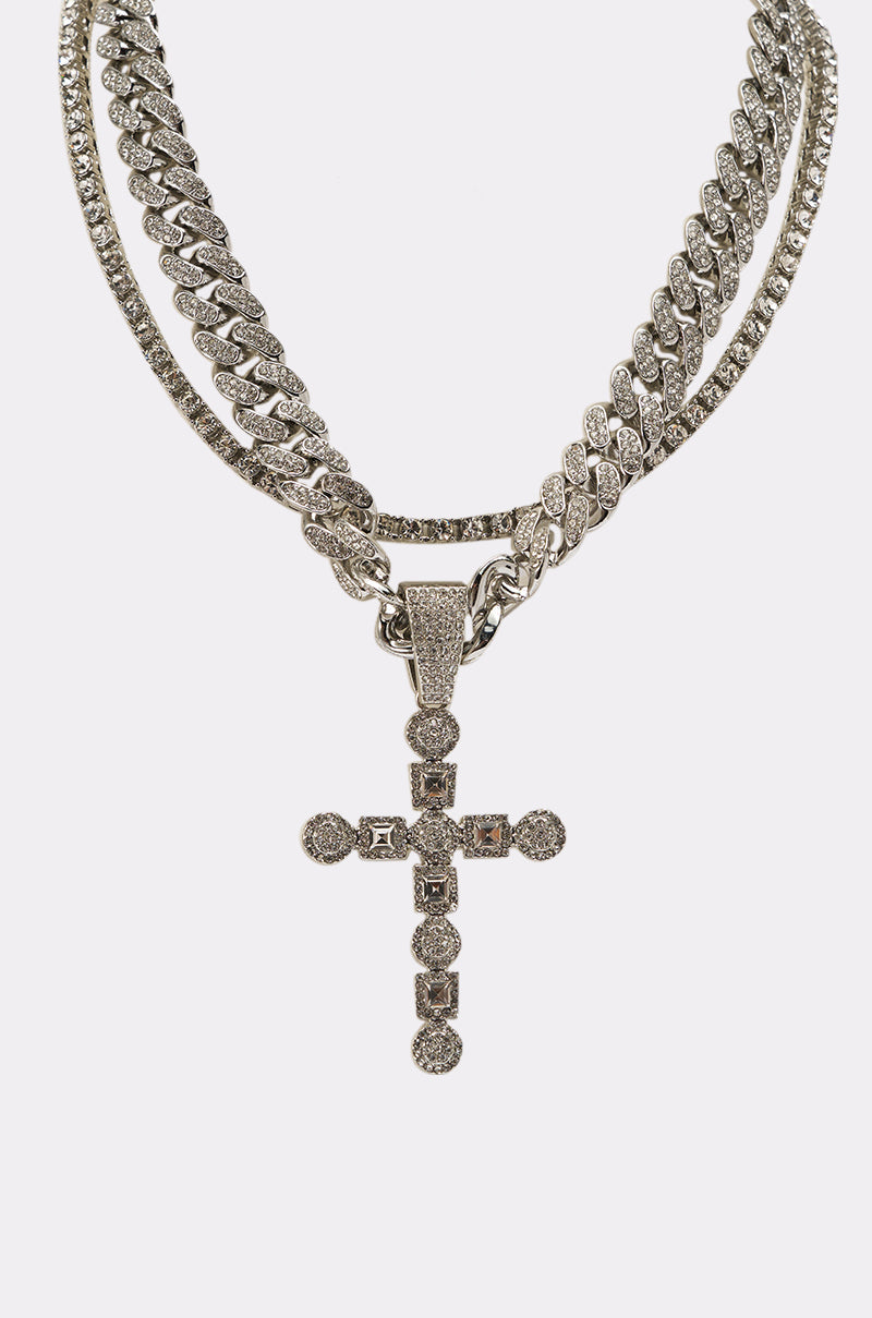 The WHAT U SAY NECKLACE is a layered set featuring a silver Cuban link chain and a large, ornate cross pendant encrusted with sparkling stones, displayed on a plain white background. 4
