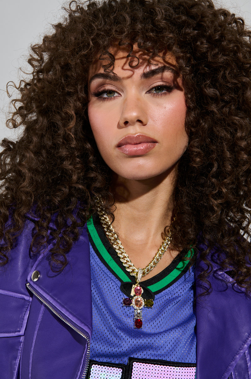 A person with curly hair wears a purple jacket over a colorful shirt, accessorized with the Dolce Vita Cross Statement Chain featuring red gems. The chunky chain link adds flair against the plain background, complementing their confident expression. 3