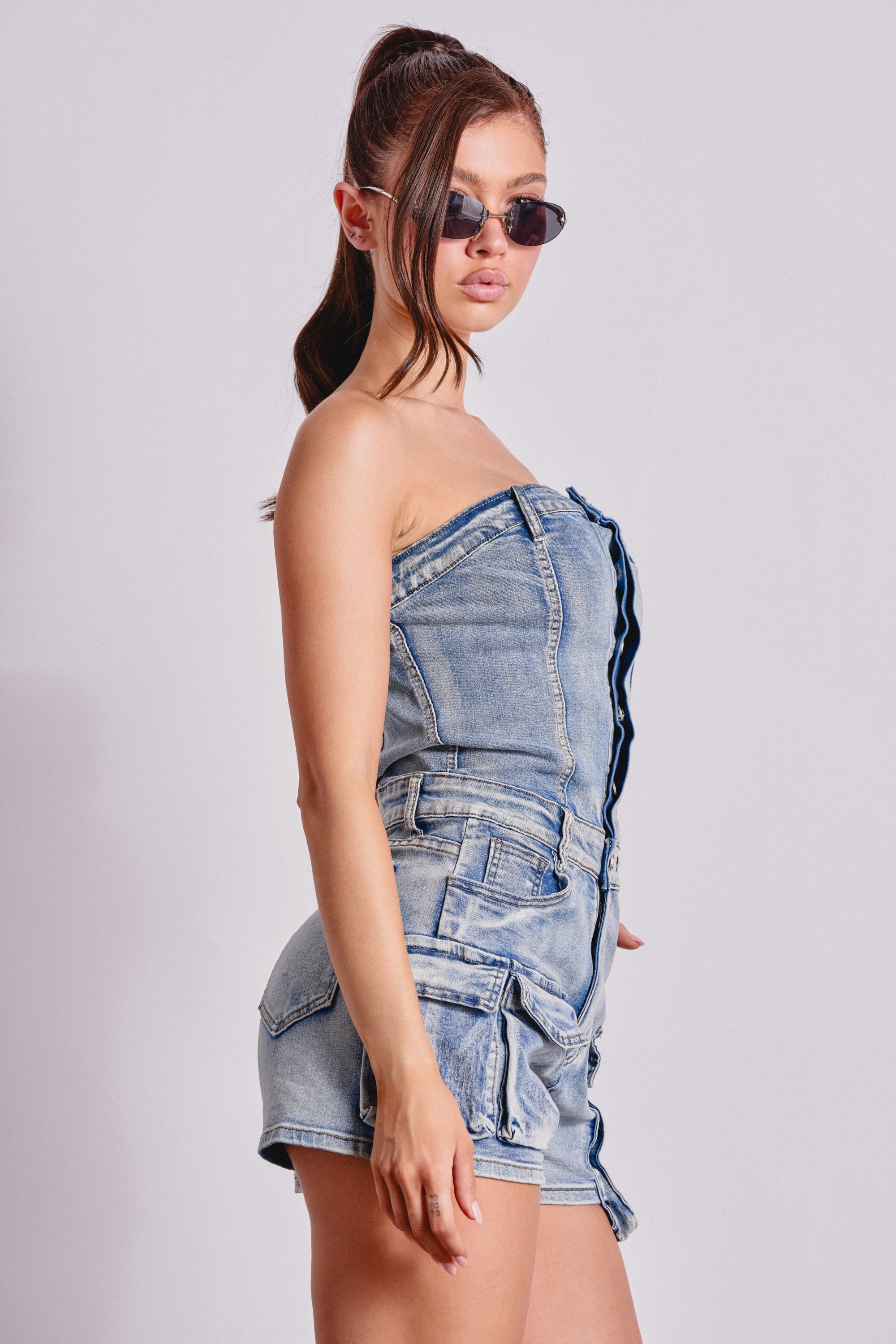 A woman models the SEE ME WIN DENIM ROMPER IN LIGHT WASH with cargo pockets and sunglasses, posing against a light background with one arm slightly bent, gazing to the side. 2