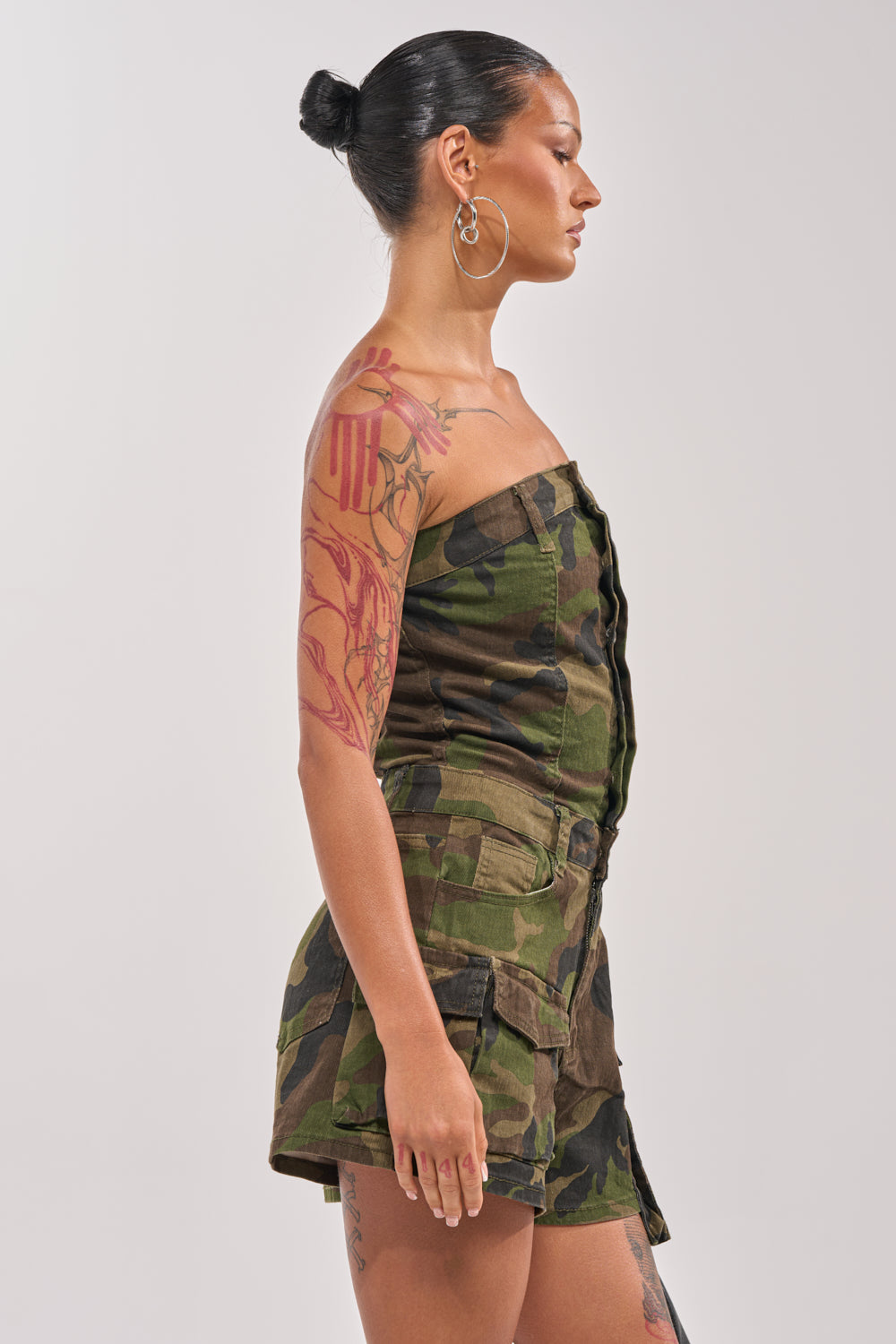 A woman with slicked-back hair and hoop earrings stands in profile, wearing the SEE ME WIN DENIM ROMPER IN CAMO. She displays colorful tattoos on her arm and leg against a plain, light background. 3
