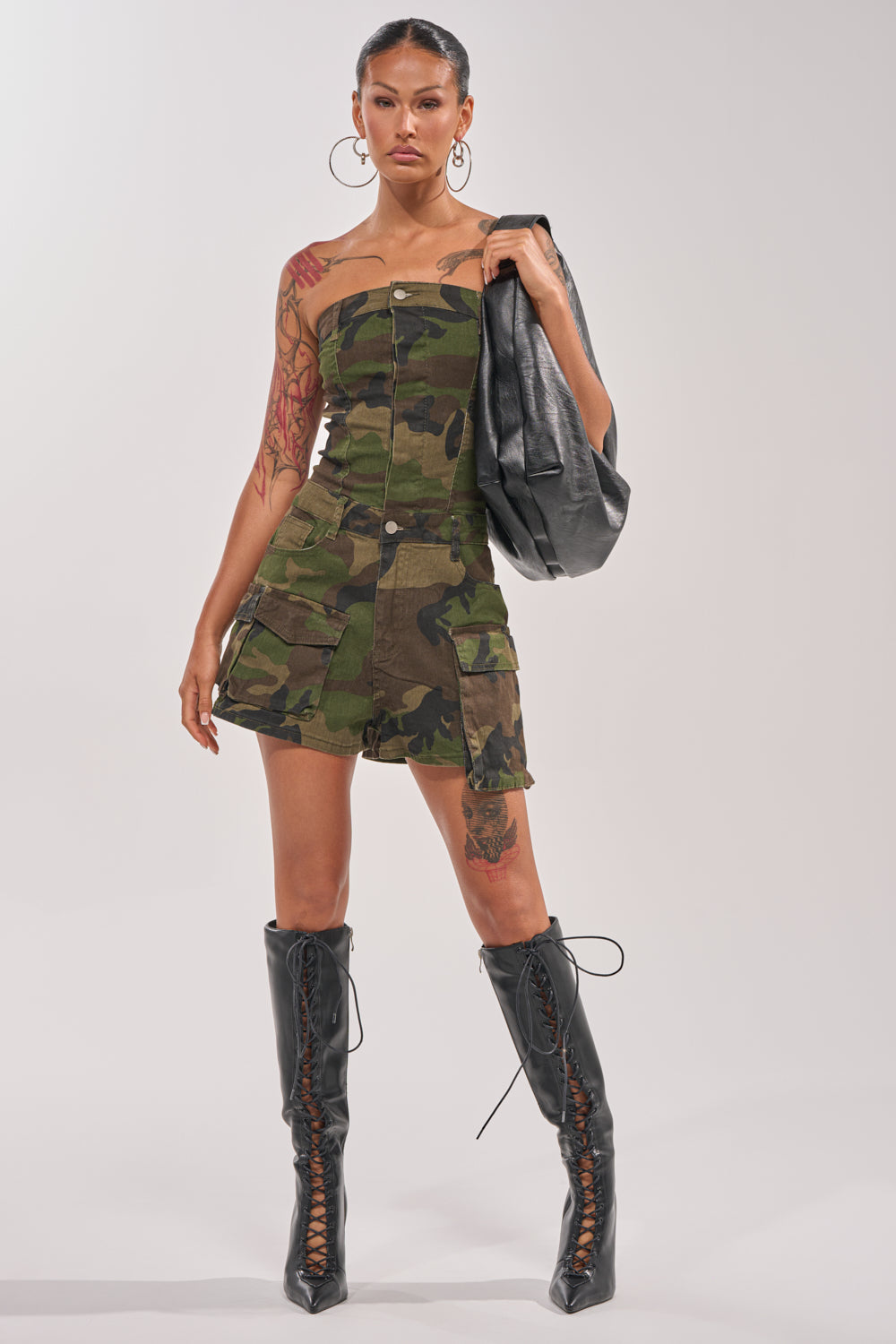 A woman stands confidently in the SEE ME WIN DENIM ROMPER IN CAMO, styled strapless with snap buttons. She pairs it with black lace-up knee-high boots, large hoop earrings, a black jacket in hand, and showcases tattoos on her arms and legs. 6