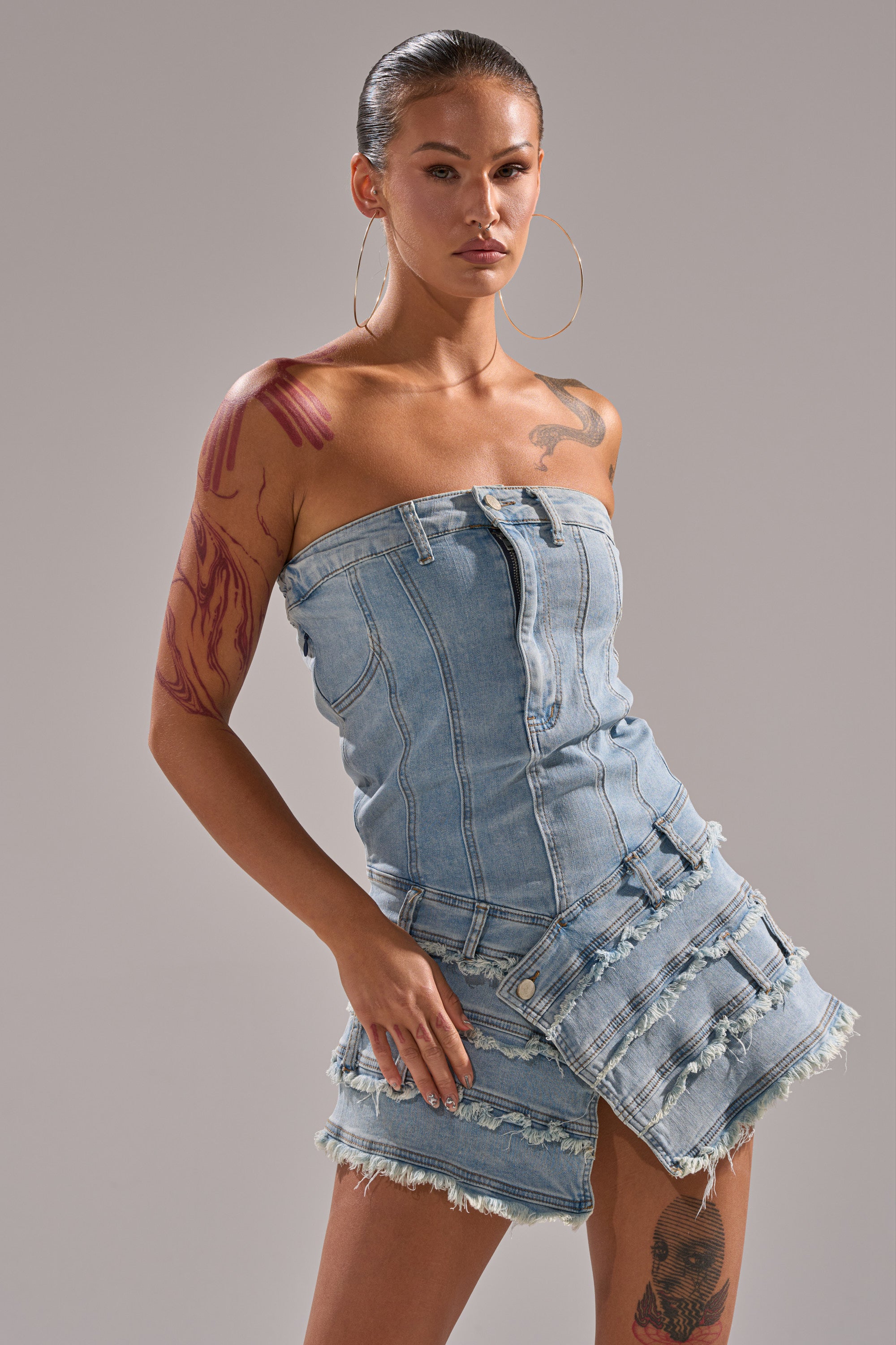 A model with large hoop earrings and tattoos showcases the TOO MUCH TO LOVE DENIM ROMPER, a strapless bustier style with frayed edges, set against a plain gray background. 2