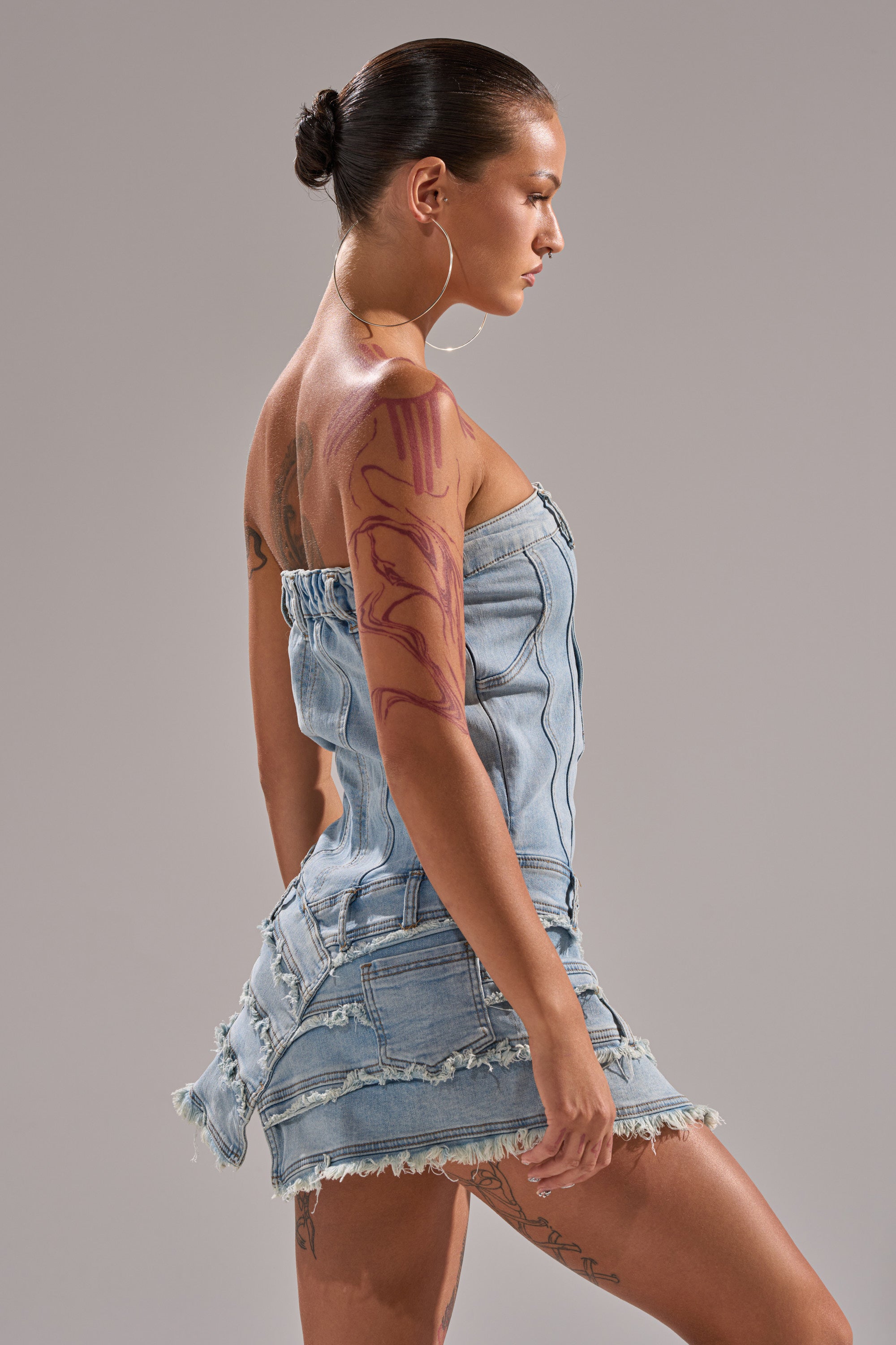 A person with slicked-back hair and large hoop earrings models the TOO MUCH TO LOVE DENIM ROMPER, a strapless, zip-up, distressed denim piece. They have visible arm tattoos and stand in profile against a neutral background. 3