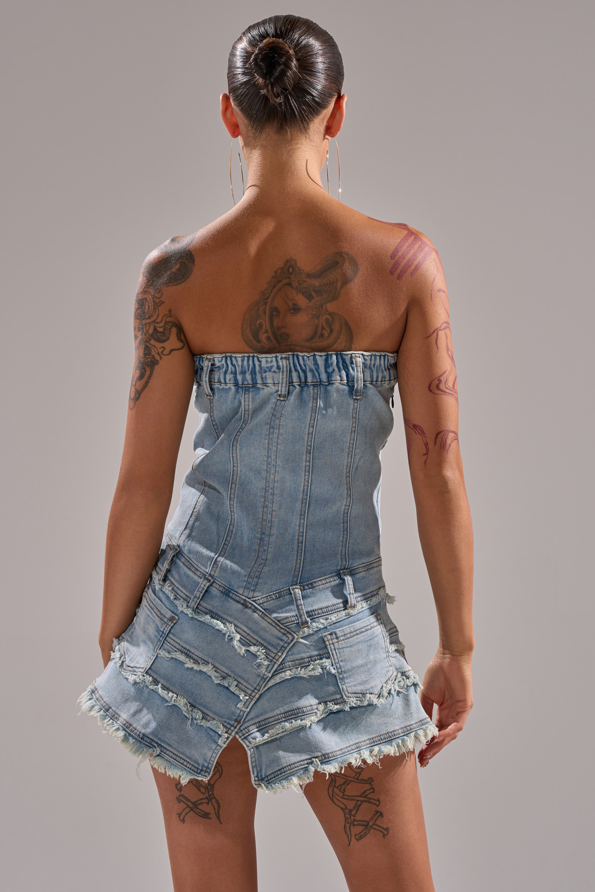 A person with tattoos, hair in a neat bun, faces away from the camera wearing the TOO MUCH TO LOVE DENIM ROMPER, a strapless piece made from upcycled jeans featuring frayed edges and visible pockets. 4