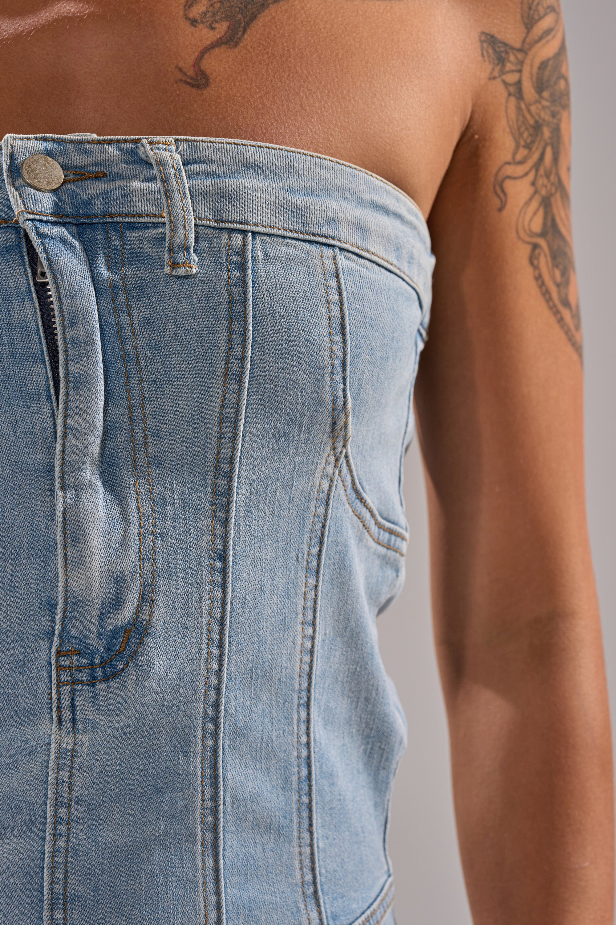 Close-up of a person wearing the TOO MUCH TO LOVE DENIM ROMPER, a strapless outfit with visible seams and button details, revealing their bare shoulder and arm with floral tattoos. 7