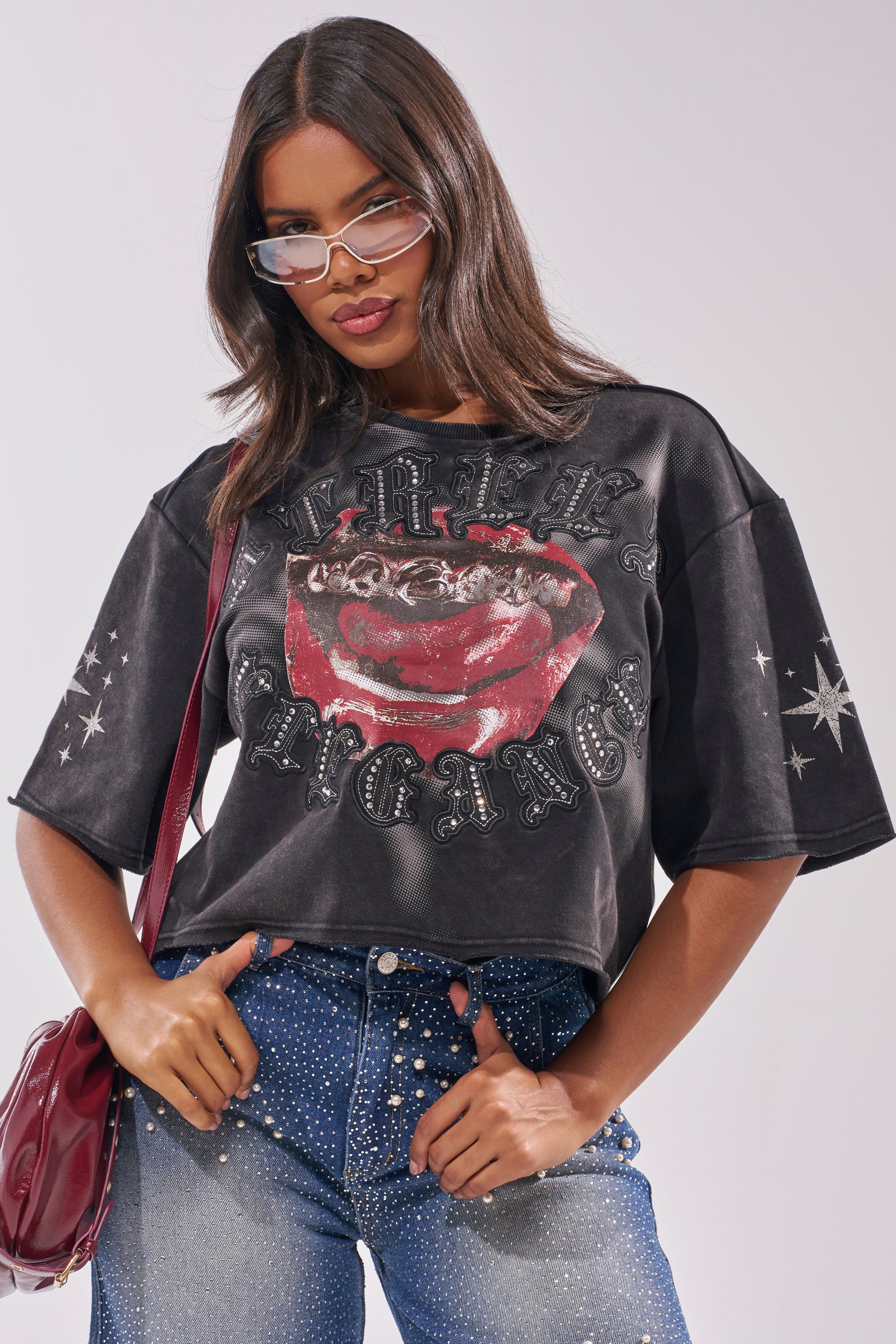 A woman exudes confidence in the STREET ELEGANCE GRAPHIC TEE, featuring a bold red lips and tongue design, paired with pearl-embellished blue jeans, chic sunglasses, and a red purse slung over her shoulder. 1