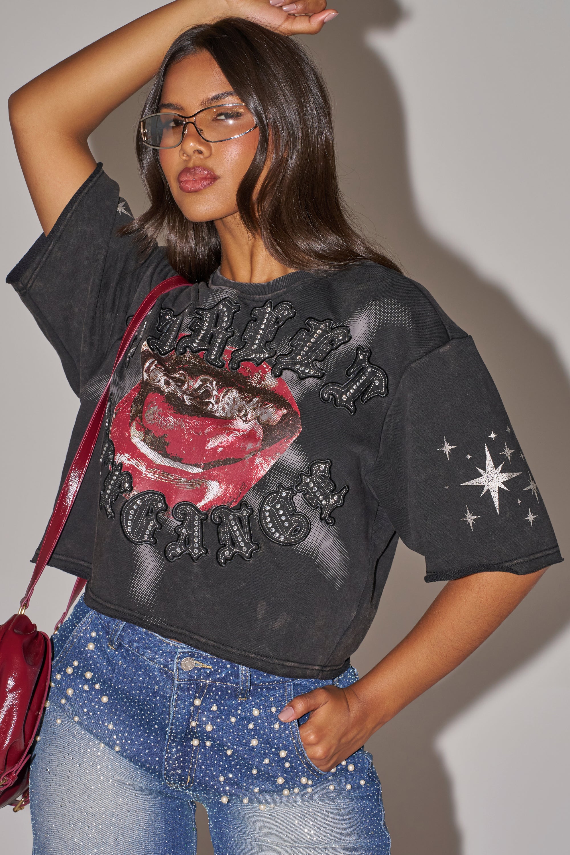 A woman exudes statement streetwear energy, wearing sunglasses and the STREET ELEGANCE GRAPHIC TEE—a black shirt with a red mouth print and "SWEET REVENGE"—paired with pearl- and rhinestone-embellished blue jeans. 2