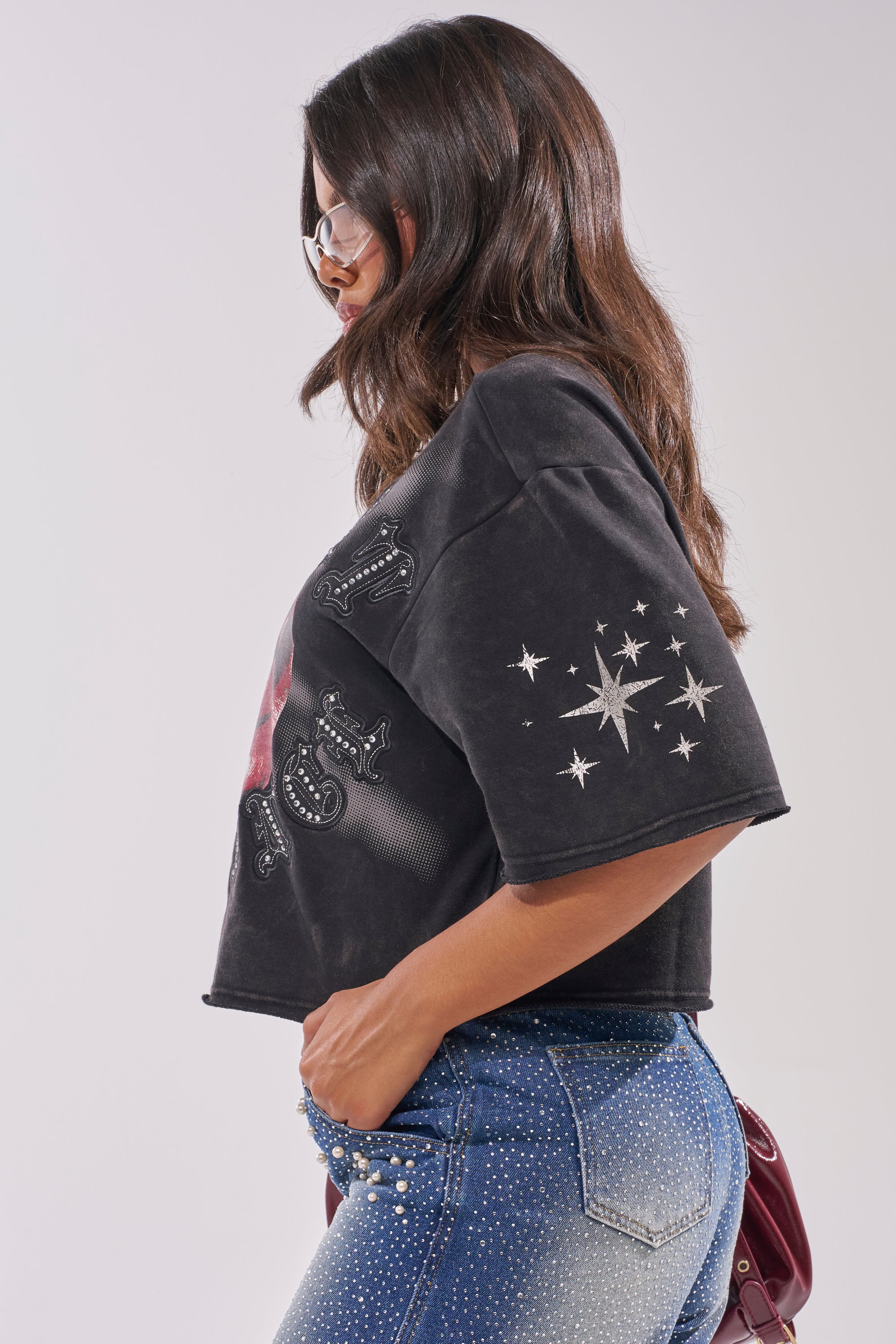 A woman with long brown hair wears sunglasses, blue jeans with white rhinestones, and the STREET ELEGANCE GRAPHIC TEE featuring star designs on the sleeve. She stands in profile against a light background, highlighting her statement streetwear style. 4