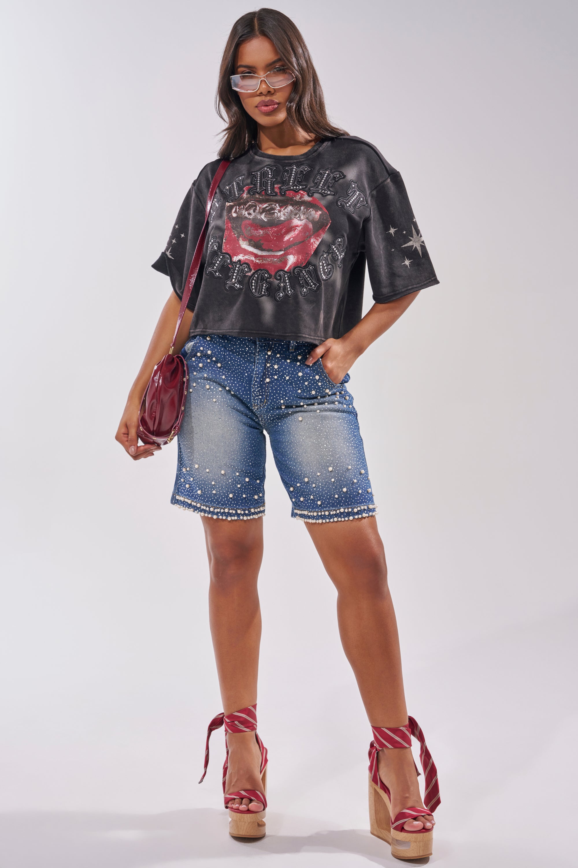A woman confidently poses against a plain backdrop wearing the STREET ELEGANCE GRAPHIC TEE, paired with studded denim Bermuda shorts, red platform heels with ankle straps, a red shoulder bag, and sunglasses. 7