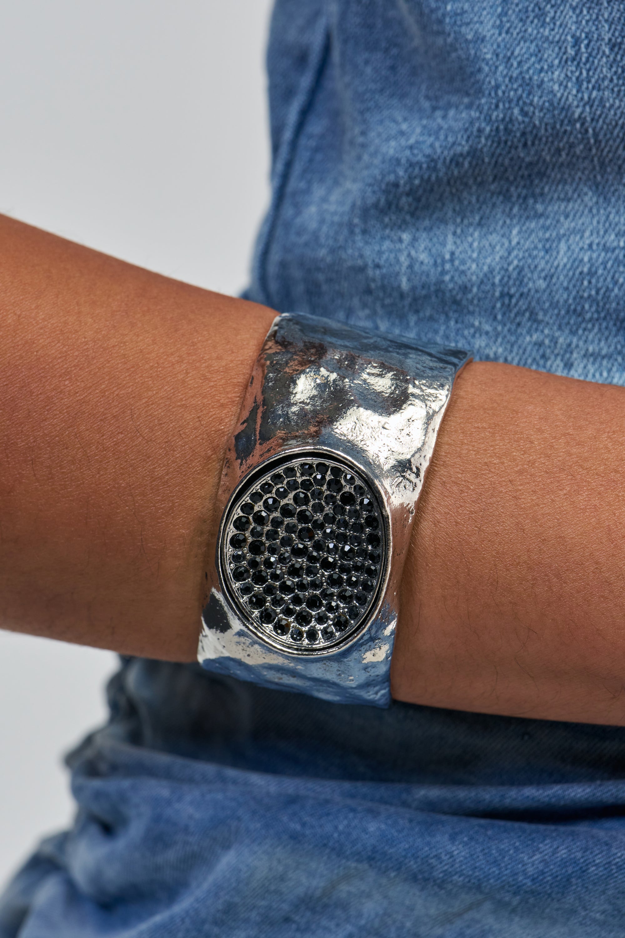 A close-up of a person wearing the MELTED METAL CUFF, a wide, hammered silver bracelet featuring a round, textured black centerpiece. This statement piece stands out boldly against the person’s blue denim clothing.
