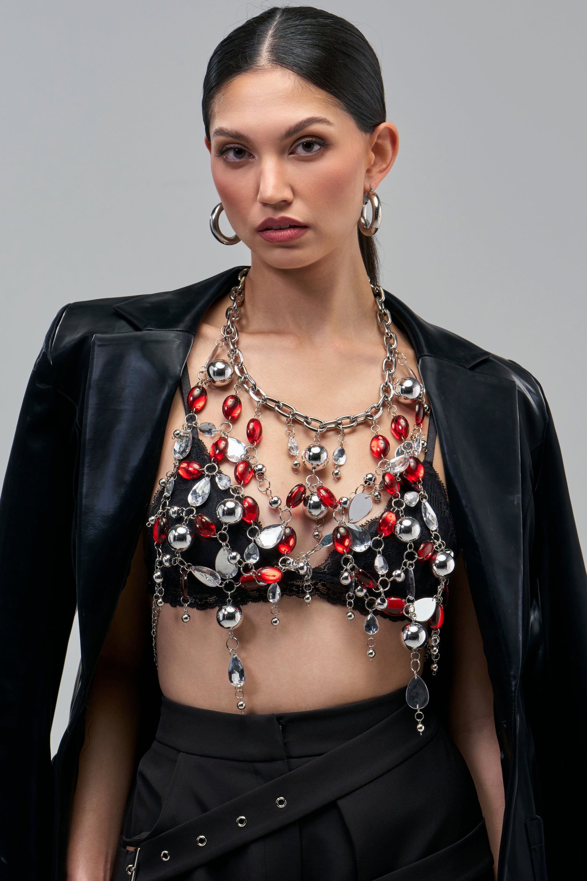 A person wears a JEWELED GODDESS BODYCHAIN with red and clear crystal teardrops over a black lace bralette and blazer.