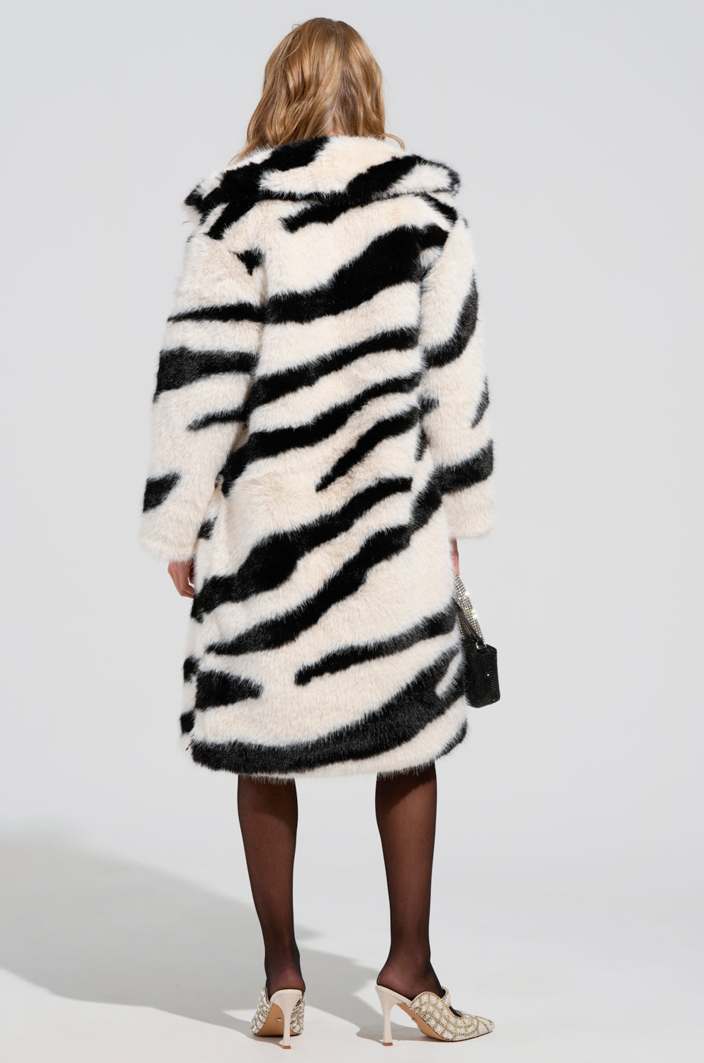 A person in an oversized BROOKFIELD FAUX FUR COAT with zebra-inspired faux fur and black tights stands facing away. They're holding a small handbag and wearing white high-heeled shoes against a plain, light-colored background.