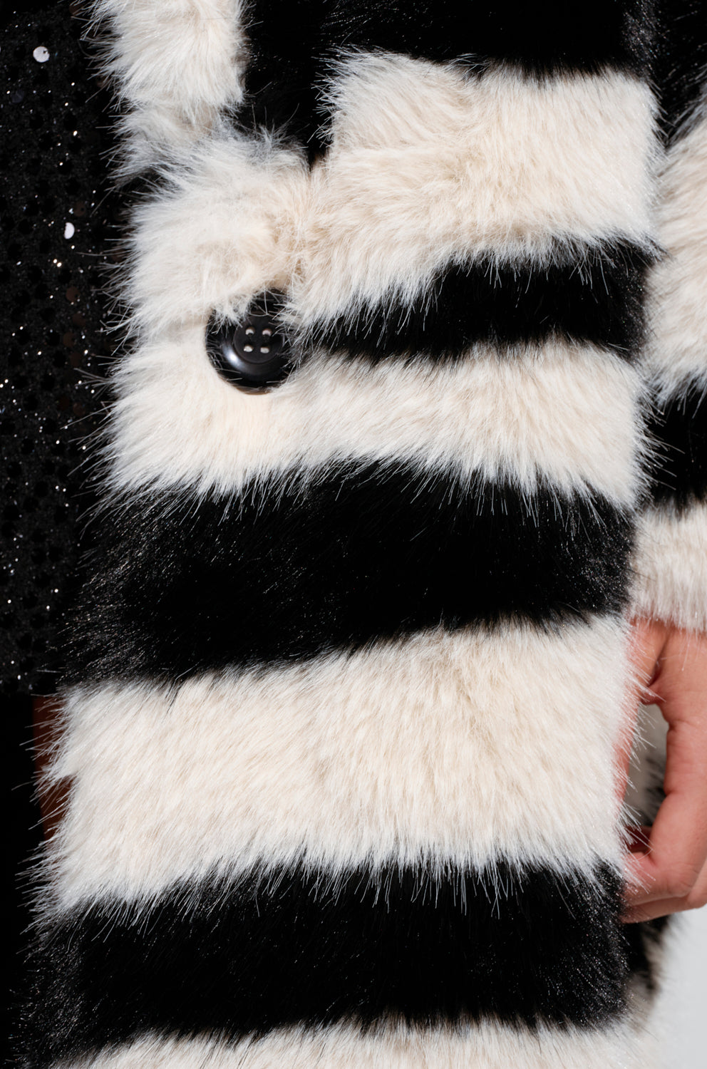 Close-up of a person wearing the BROOKFIELD FAUX FUR COAT, an oversized black and white zebra-inspired design with large buttons. A hand rests near the coat on the right side, set against a hint of black sequined fabric in the background.