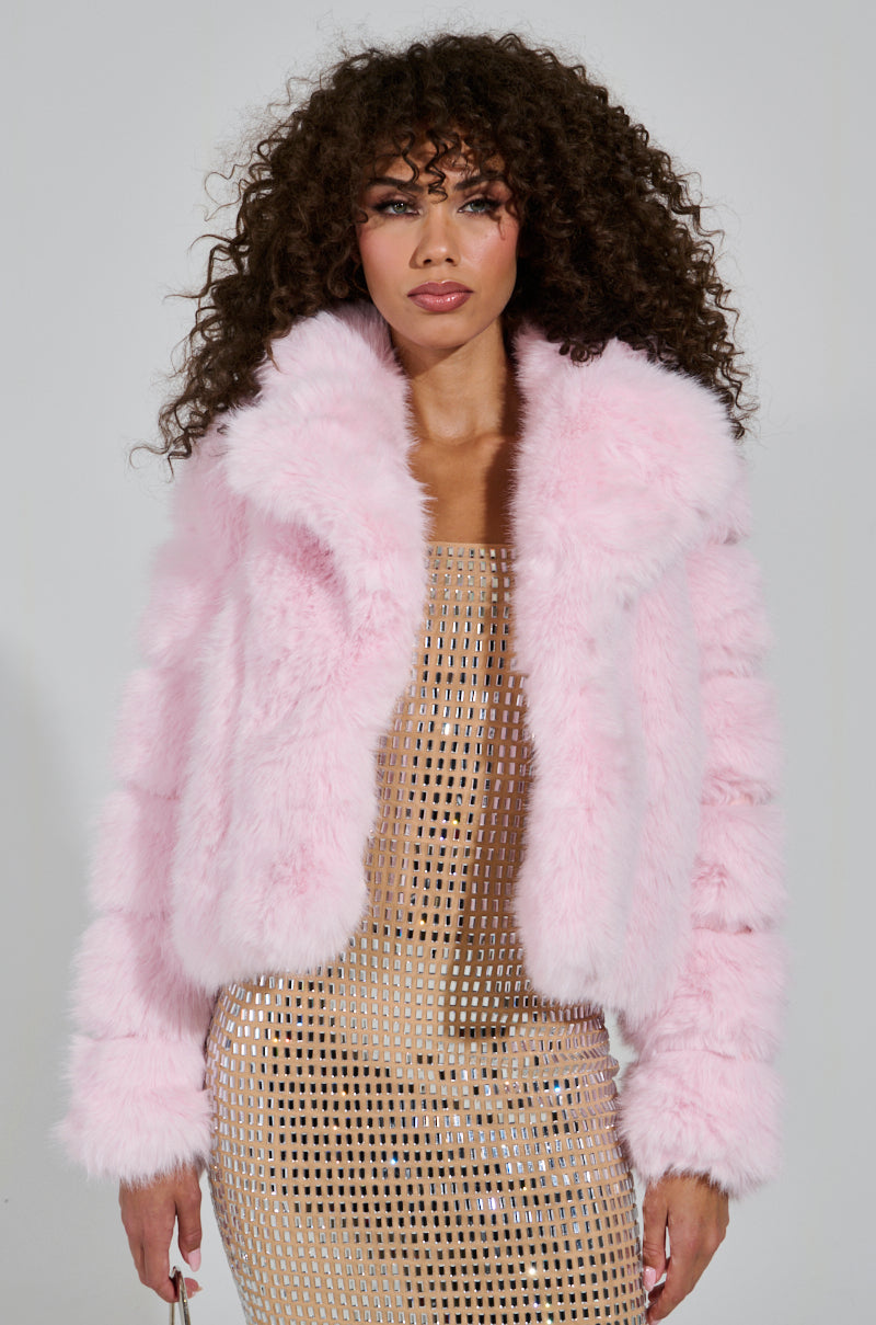 A person with curly hair confidently gazes at the camera, wearing the BERRIES AND CREAM FAUX FUR JACKET featuring an oversized lapel collar layered over a shimmering gold dress. The cozy ensemble stands out brilliantly against the plain white background.