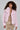 A person with curly hair is dressed in a cozy ensemble, showcasing the BERRIES AND CREAM FAUX FUR JACKET in a fluffy pink hue draped over a sparkly gold dress. They stylishly hold a small handbag while posing against a plain background.