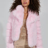 A person with curly hair is dressed in a cozy ensemble, showcasing the BERRIES AND CREAM FAUX FUR JACKET in a fluffy pink hue draped over a sparkly gold dress. They stylishly hold a small handbag while posing against a plain background.