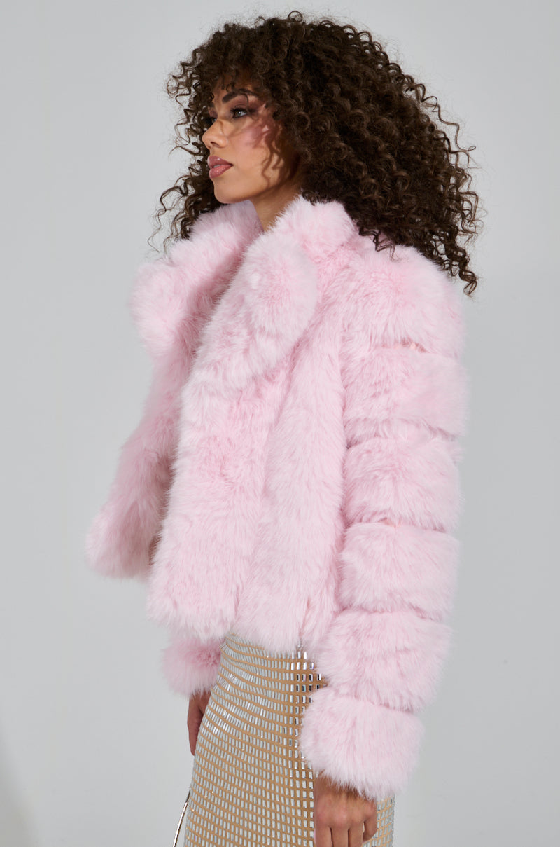 A person with curly hair is wearing the BERRIES AND CREAM FAUX FUR JACKET, featuring a fluffy pink design and an oversized lapel collar. They pair it with a sparkly skirt while standing sideways to the camera, gazing forward against a plain grey background, creating a cozy ensemble.