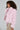 A person with curly hair is wearing the BERRIES AND CREAM FAUX FUR JACKET, featuring a fluffy pink design and an oversized lapel collar. They pair it with a sparkly skirt while standing sideways to the camera, gazing forward against a plain grey background, creating a cozy ensemble.