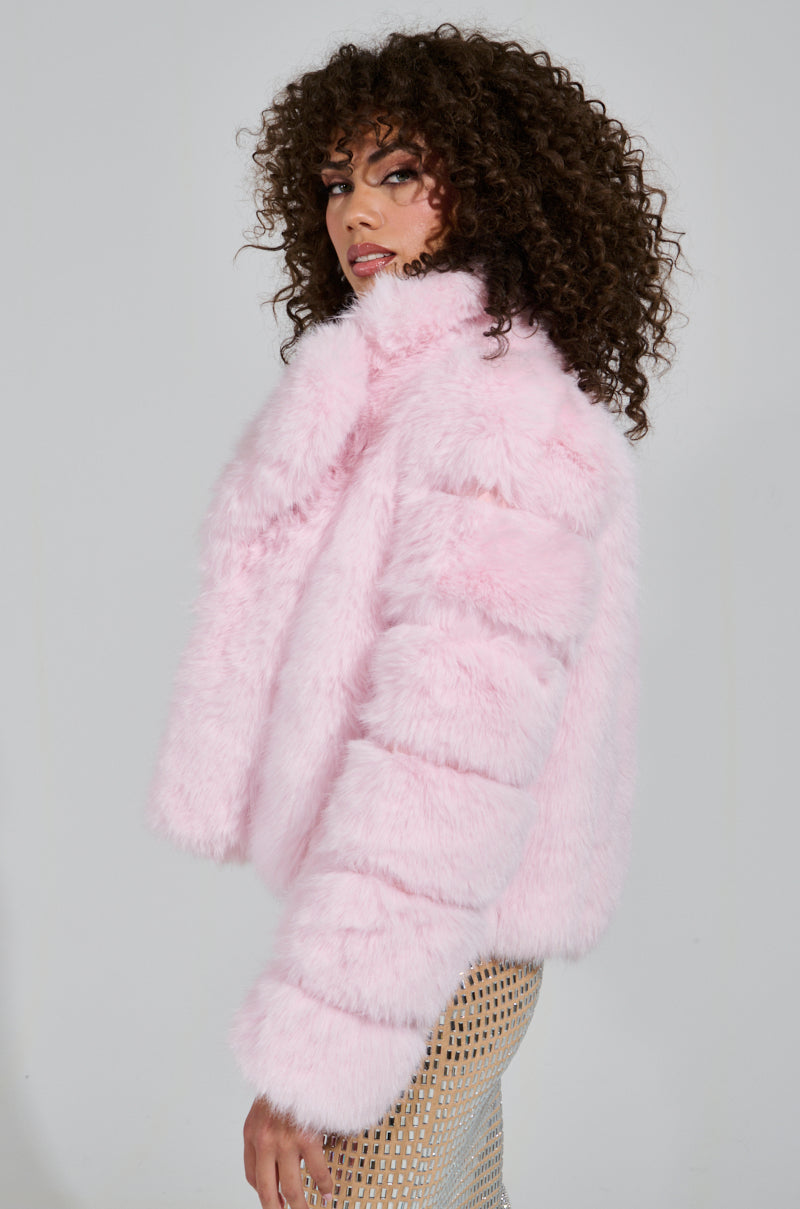 Wearing the BERRIES AND CREAM FAUX FUR JACKET with its oversized lapel collar, a person with curly hair pairs it effortlessly with a patterned skirt, glancing over their shoulder against a plain background.