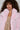 A woman with curly hair is dressed warmly in the Berries and Cream Faux Fur Jacket, which boasts a light pink hue and an oversized lapel collar. She wears it over a dress embellished with metallic squares, looking straight into the camera against a plain background.