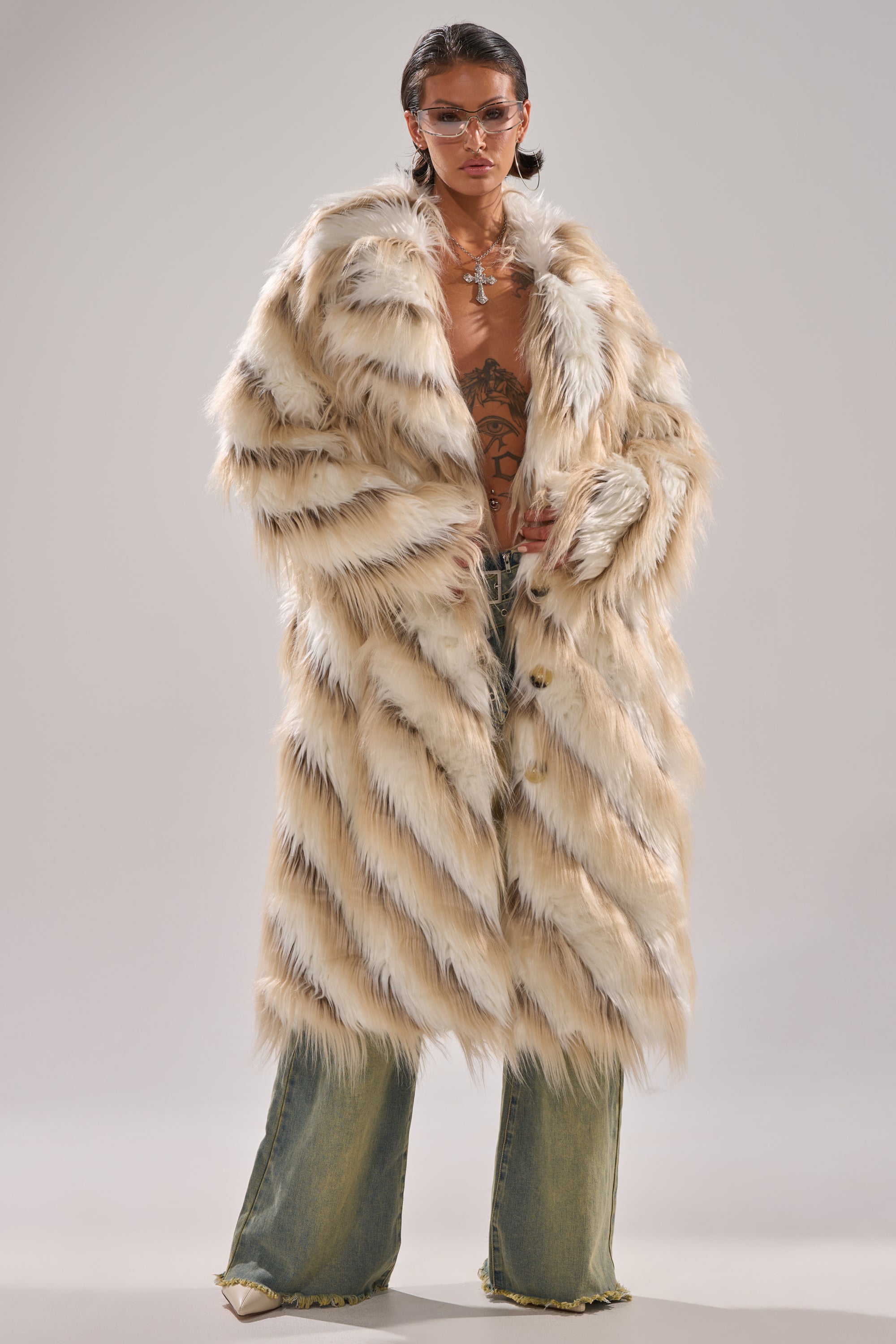 A person in oversized sunglasses, loose ripped jeans, jewelry, and the HARLOW PATCHWORK FAUX FUR coat with a longline silhouette and visible chest tattoos stands against a plain background.