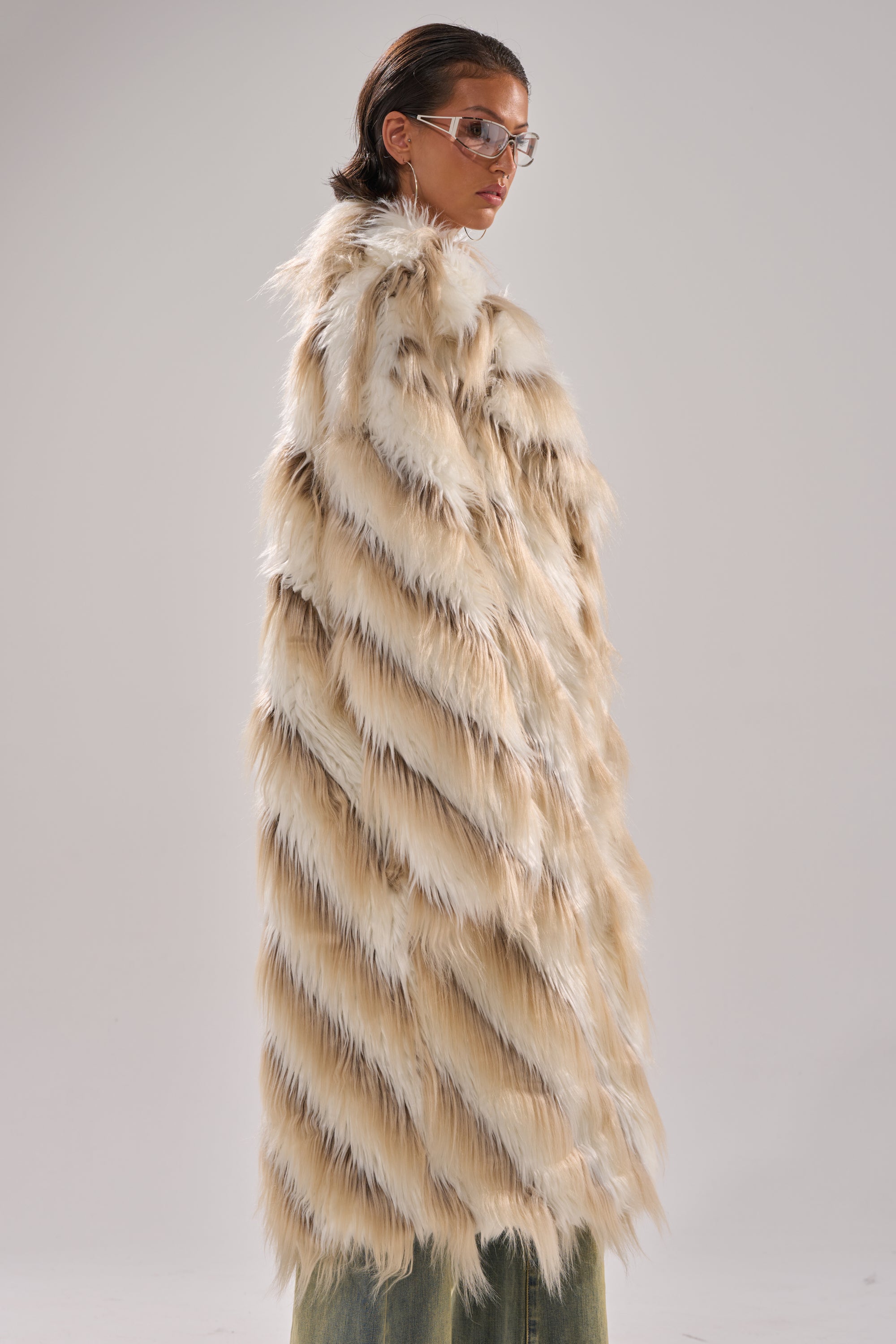 A person wearing clear glasses and the HARLOW PATCHWORK FAUX FUR coat with a longline silhouette stands facing away from the camera against a plain light background.