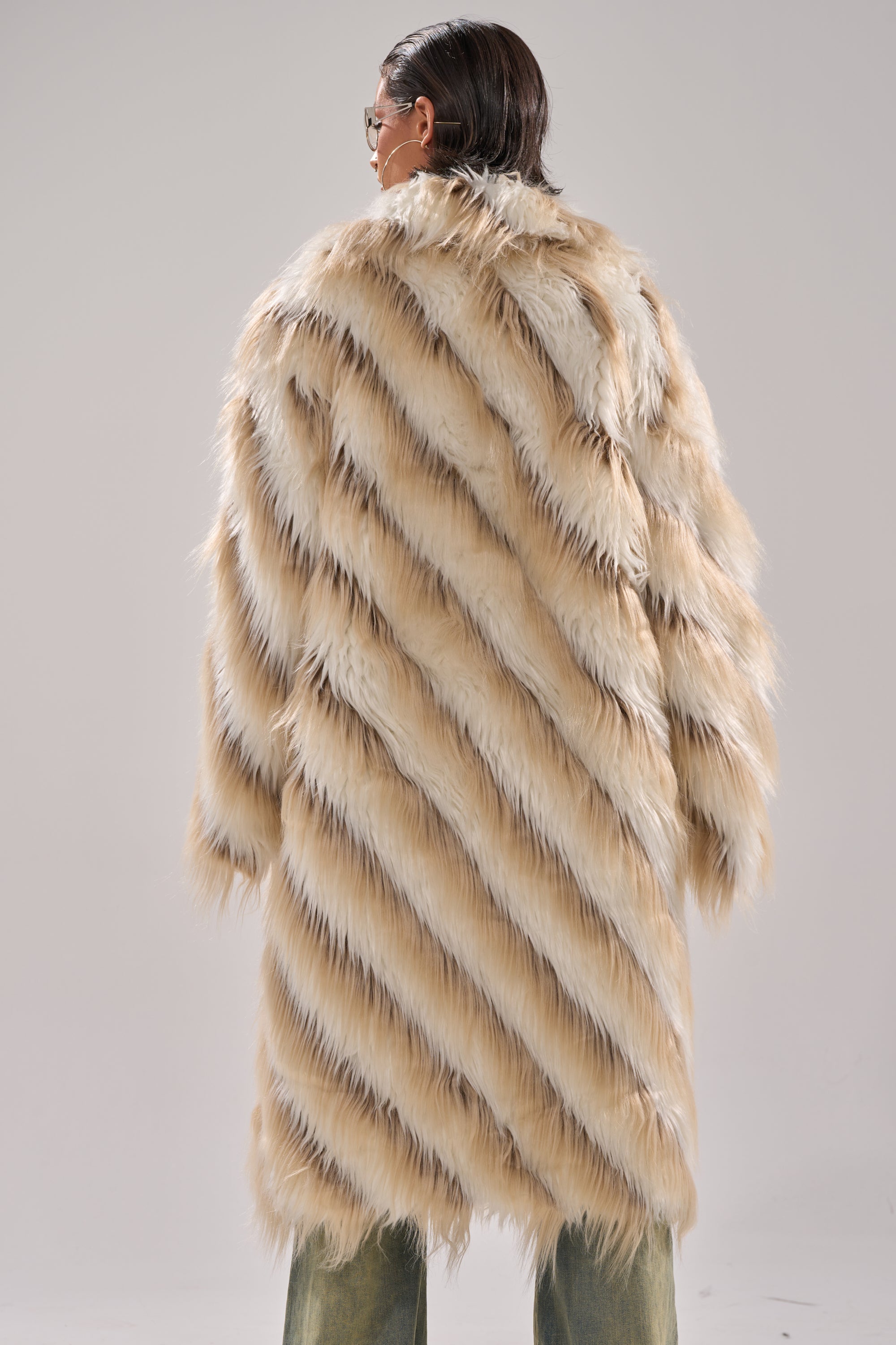 A person with short dark hair, seen from behind, wears the HARLOW PATCHWORK FAUX FUR coat in beige and white with diagonal stripes and a longline silhouette, set against a plain light gray background.