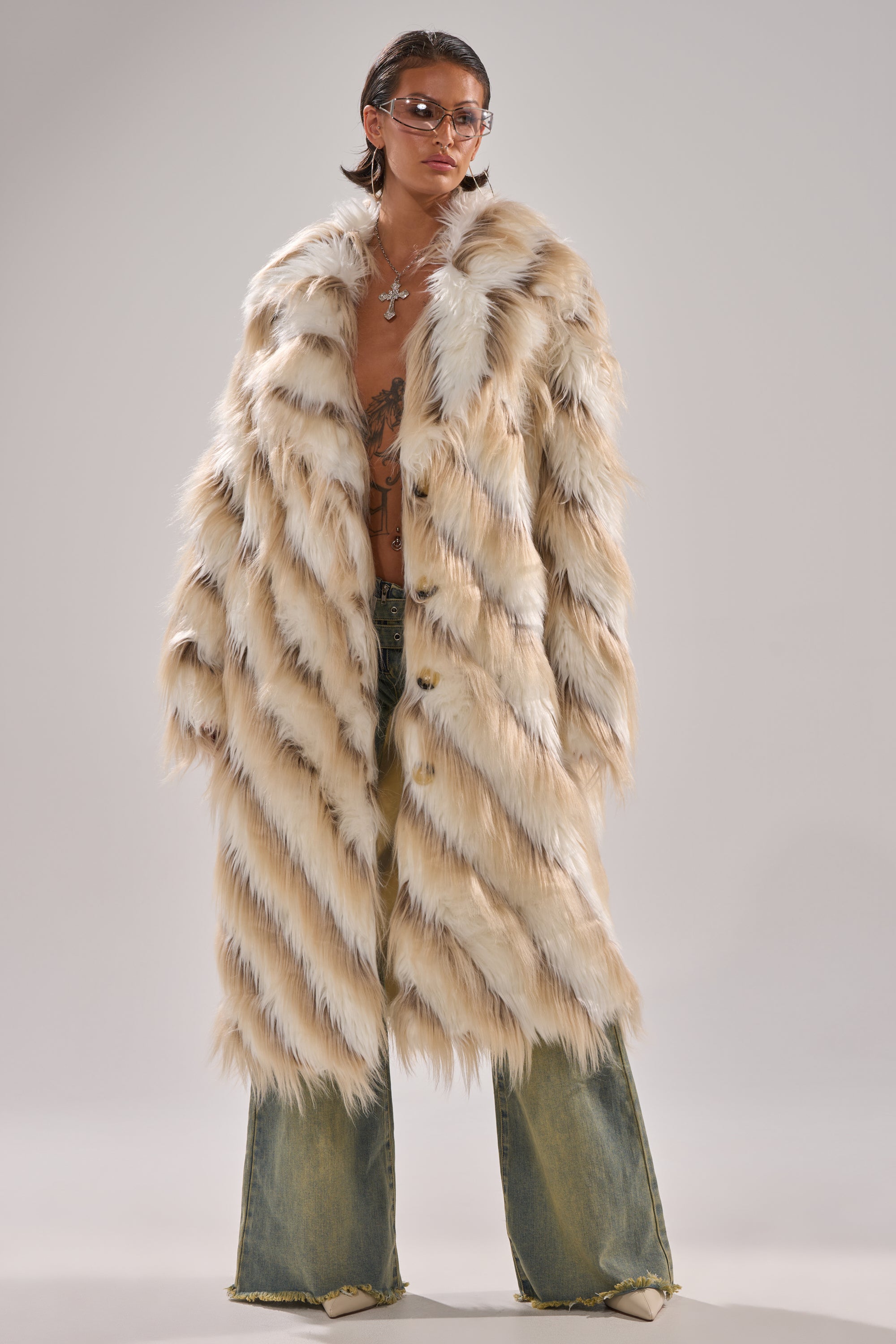 A person wears the HARLOW PATCHWORK FAUX FUR coat in cream and brown, paired with light-wash wide-leg jeans, sunglasses, and necklaces, standing confidently against a plain background.