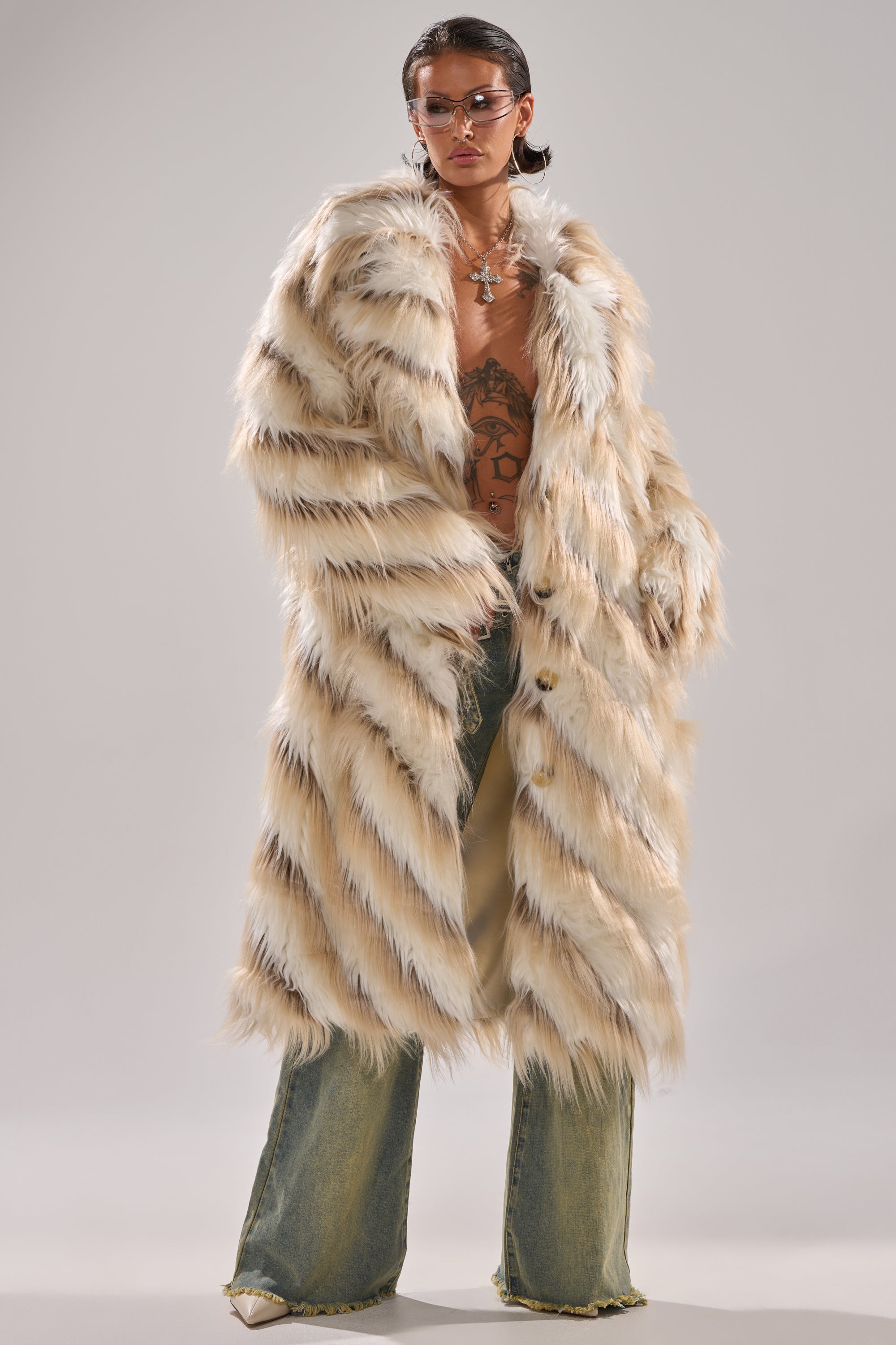 Wearing sunglasses and the HARLOW PATCHWORK FAUX FUR coat with black and white stripes, loose jeans, and displaying chest tattoos, someone poses confidently against a plain background.