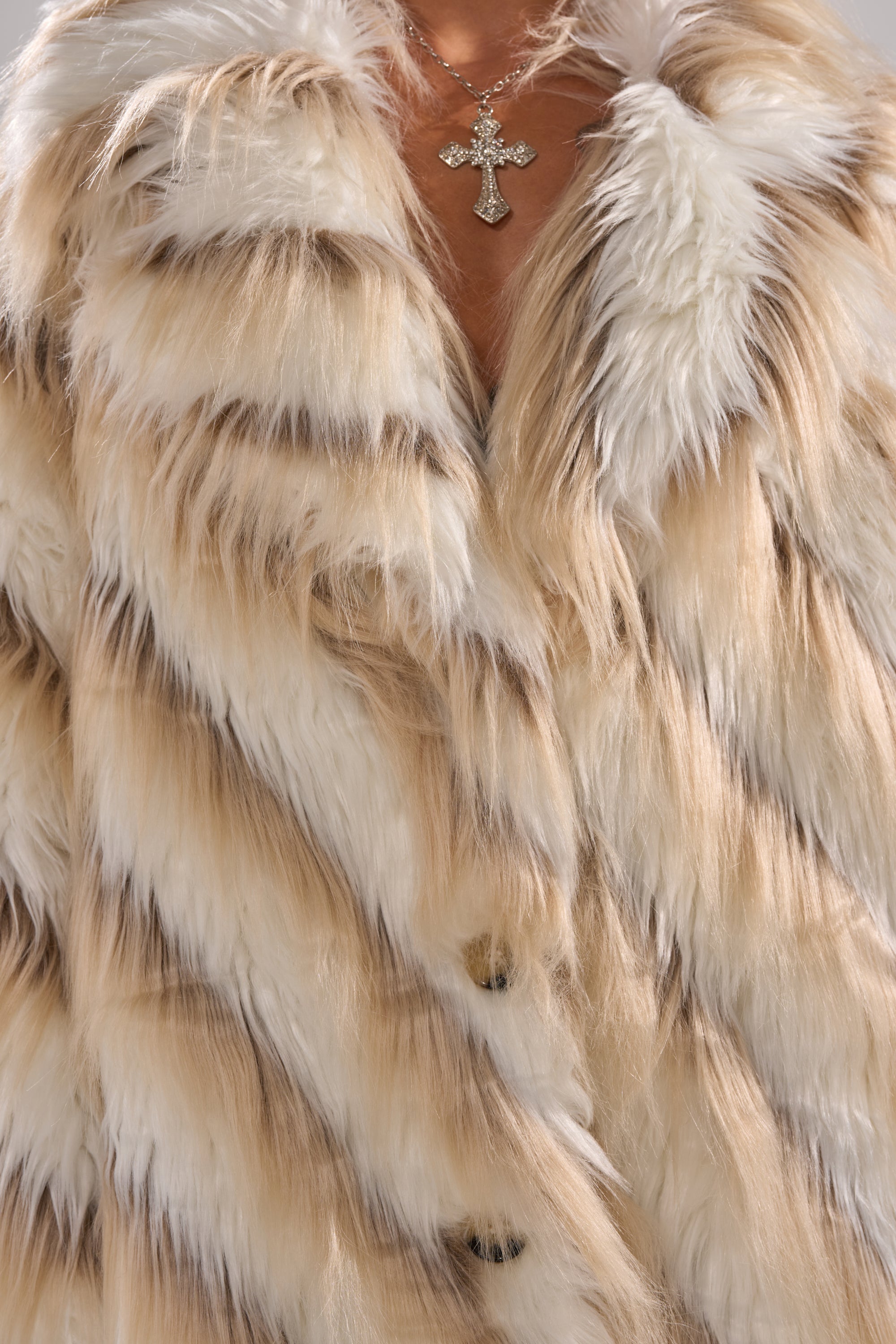 Close-up of a person wearing the HARLOW PATCHWORK FAUX FUR—an oversized striped coat in beige, white, and brown—with a cross pendant necklace. Face not visible.